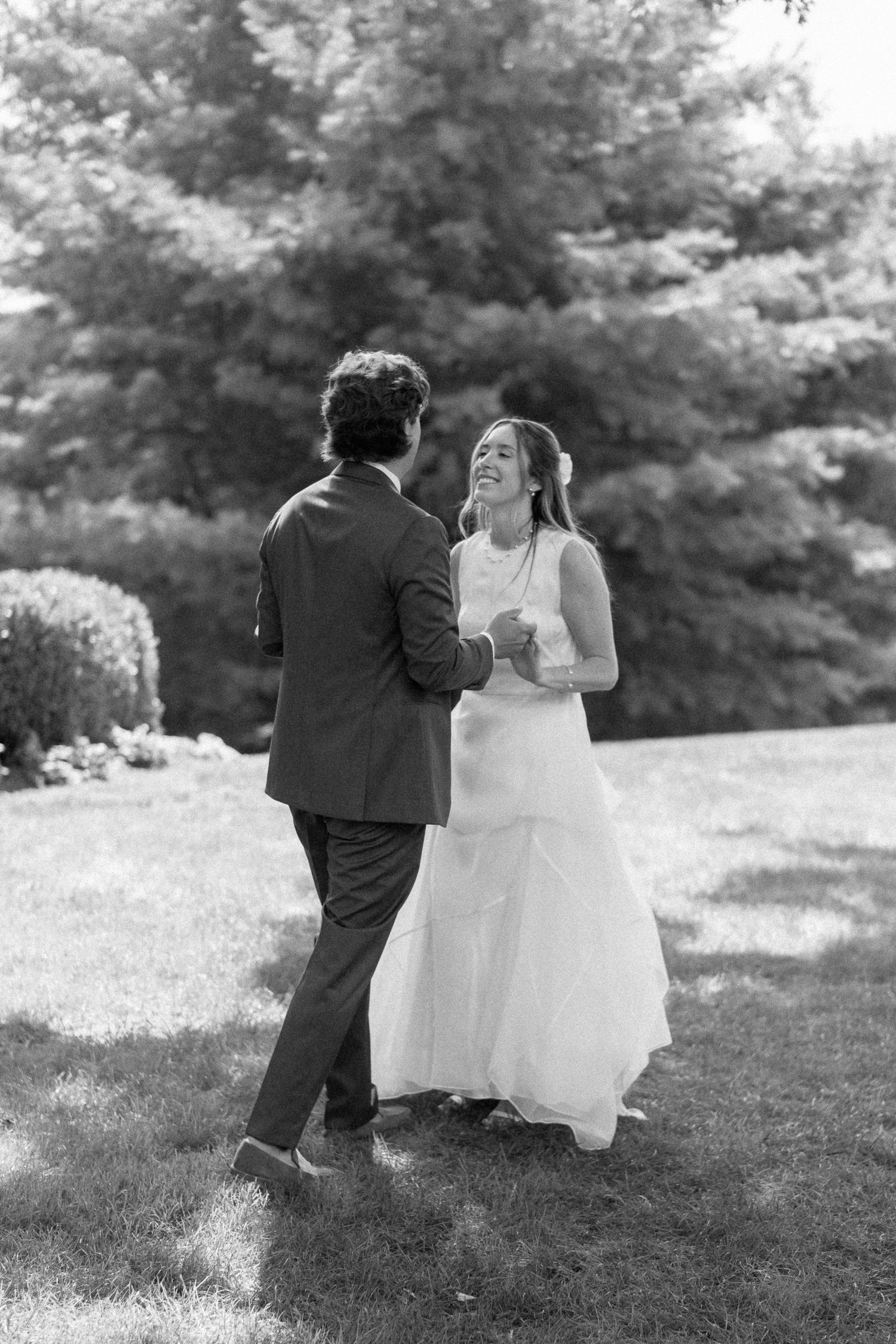 Black and white image of a joyful bride laughing while dancing with her groom on a sunny lawn.
