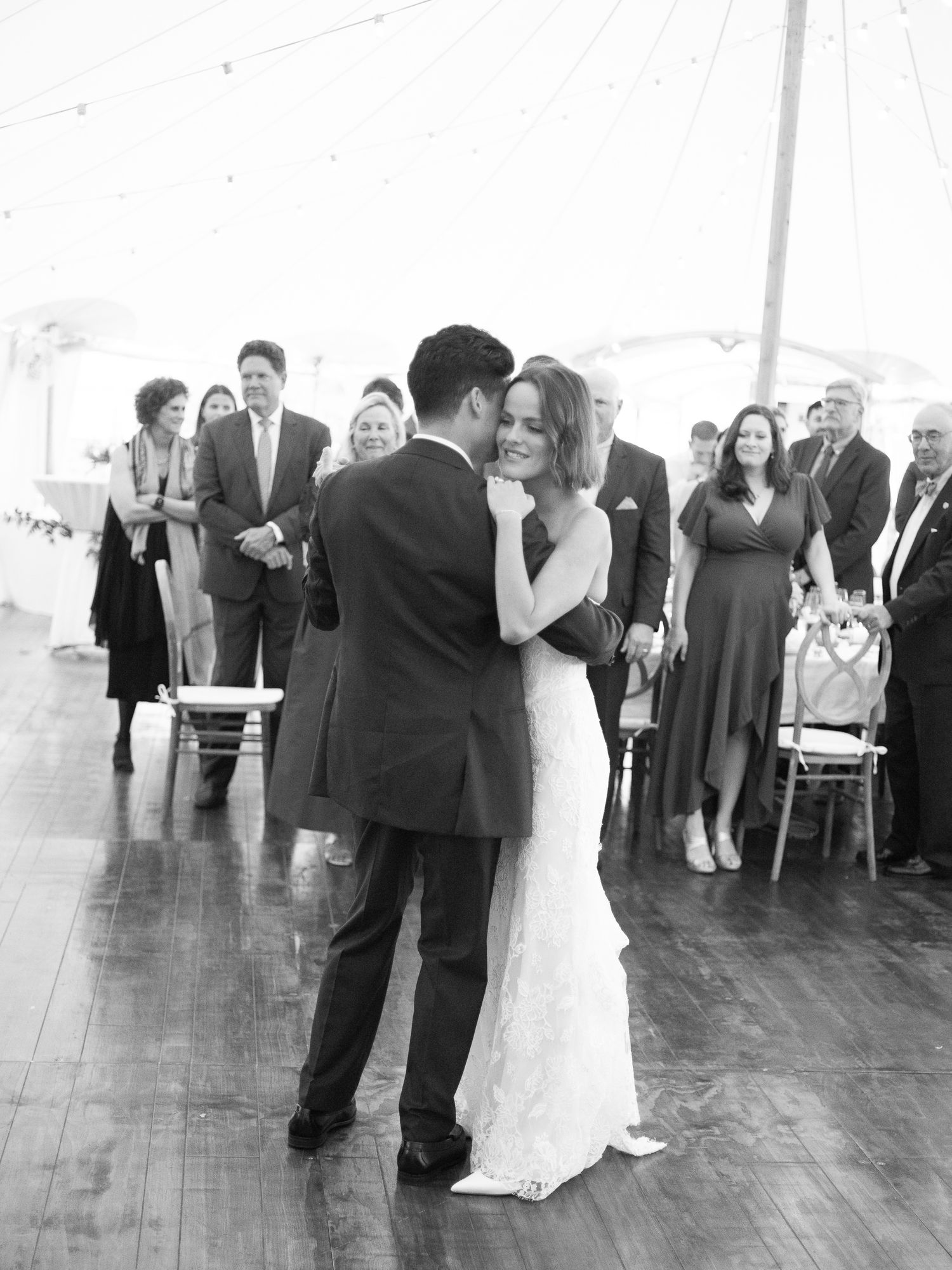 Bride and groom share romantic first dance under sailcloth tent with wedding guests watching.