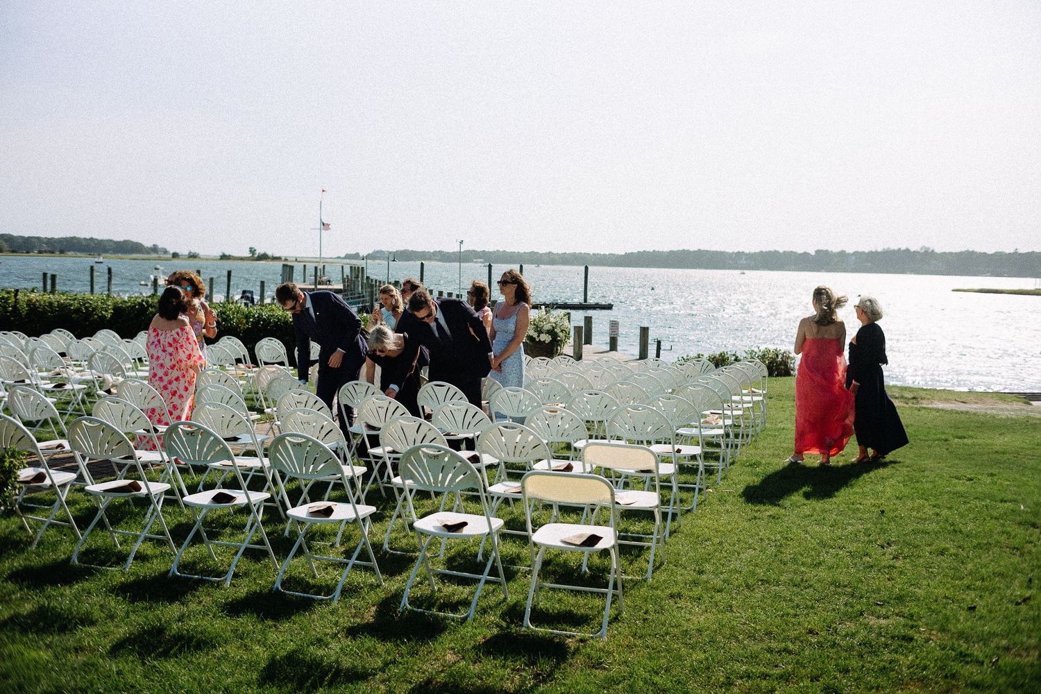 Outdoor waterfront wedding ceremony setup with white folding chairs on green lawn, guests mingling near a dock on a sunny day.
