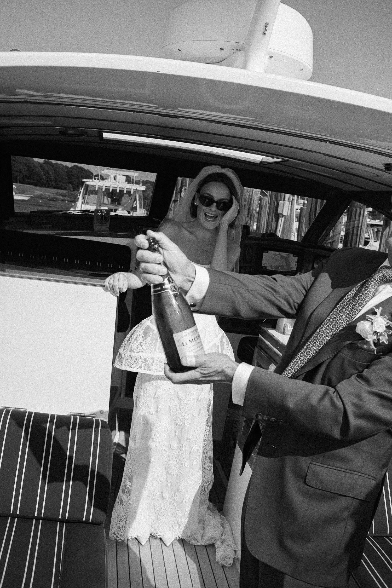 Black and white photo of a bride and groom opening champagne on a luxury yacht on their wedding day.