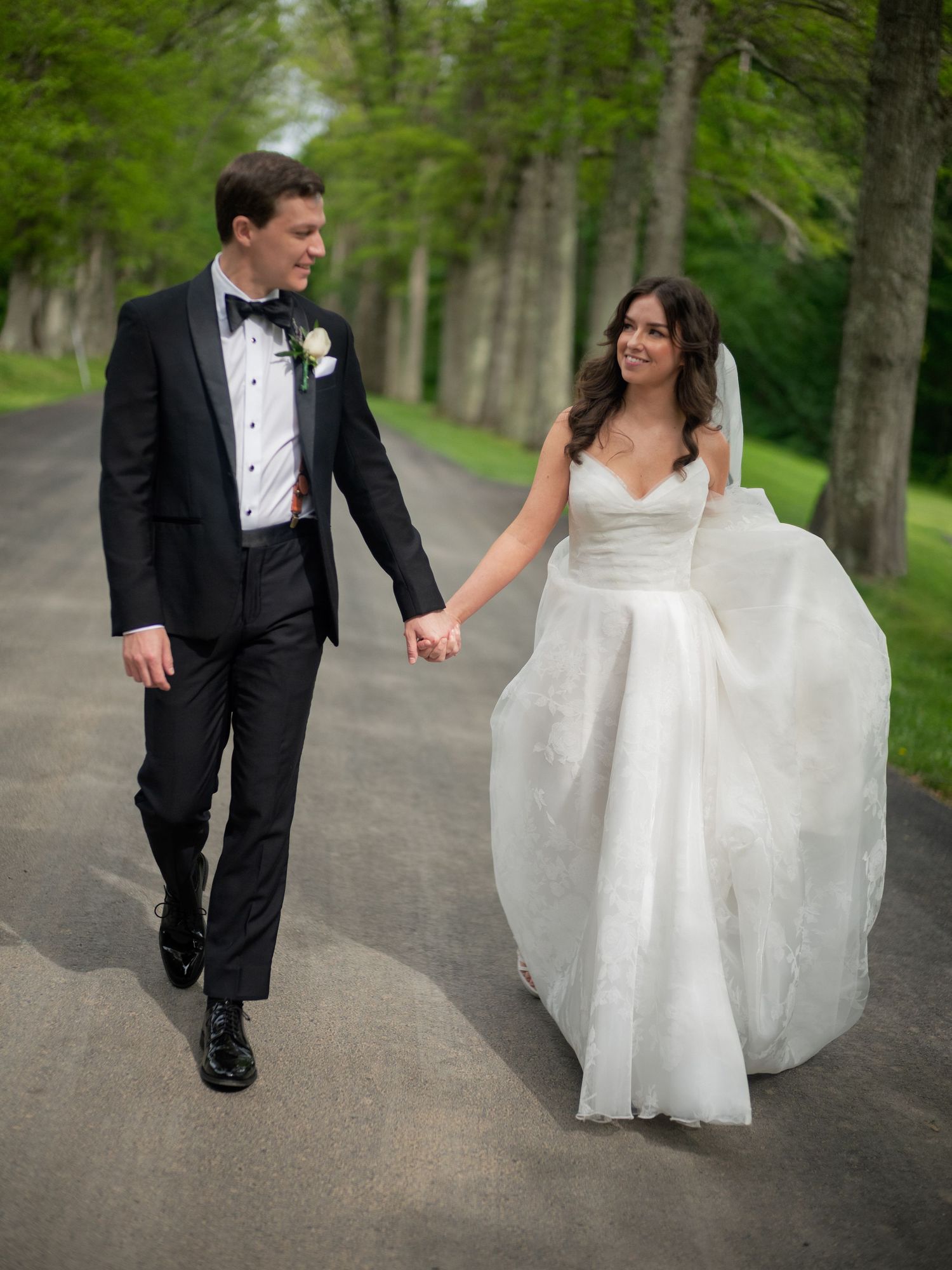 Bride and groom walk hand in hand down a tree-lined path surrounded by lush green foliage.