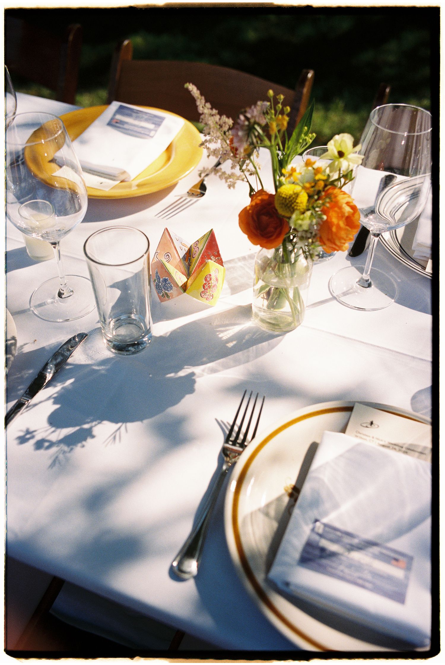 Elegantly set outdoor wedding reception table with floral centerpiece, yellow plates, and glassware.