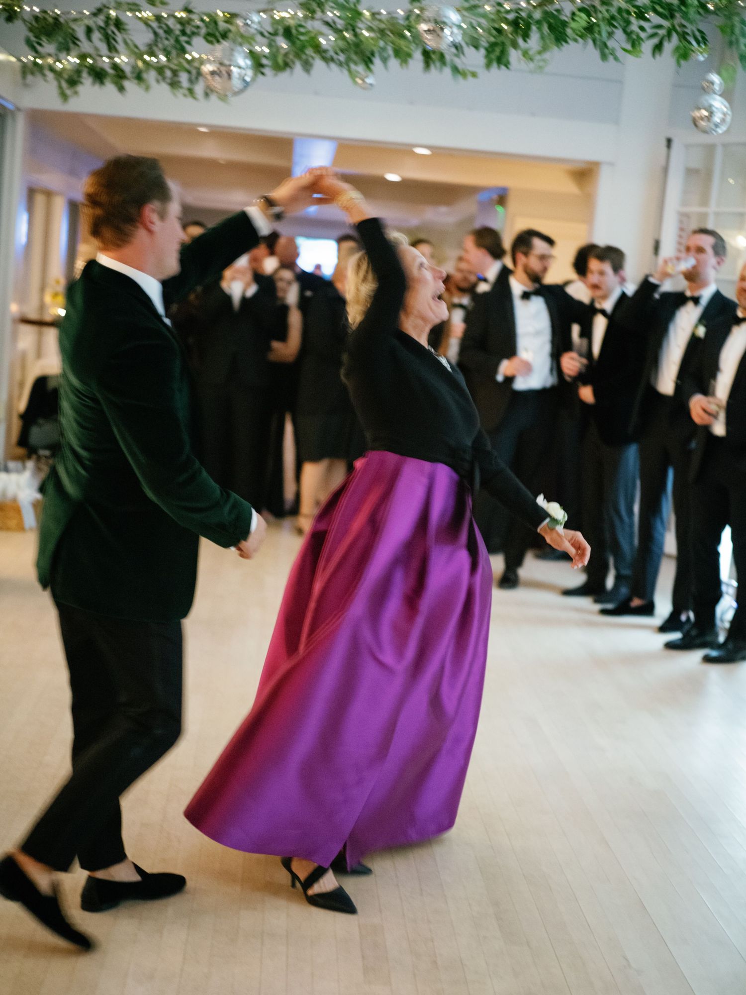 Guests dancing joyfully at wedding reception, woman in purple skirt twirling with man in green velvet jacket.