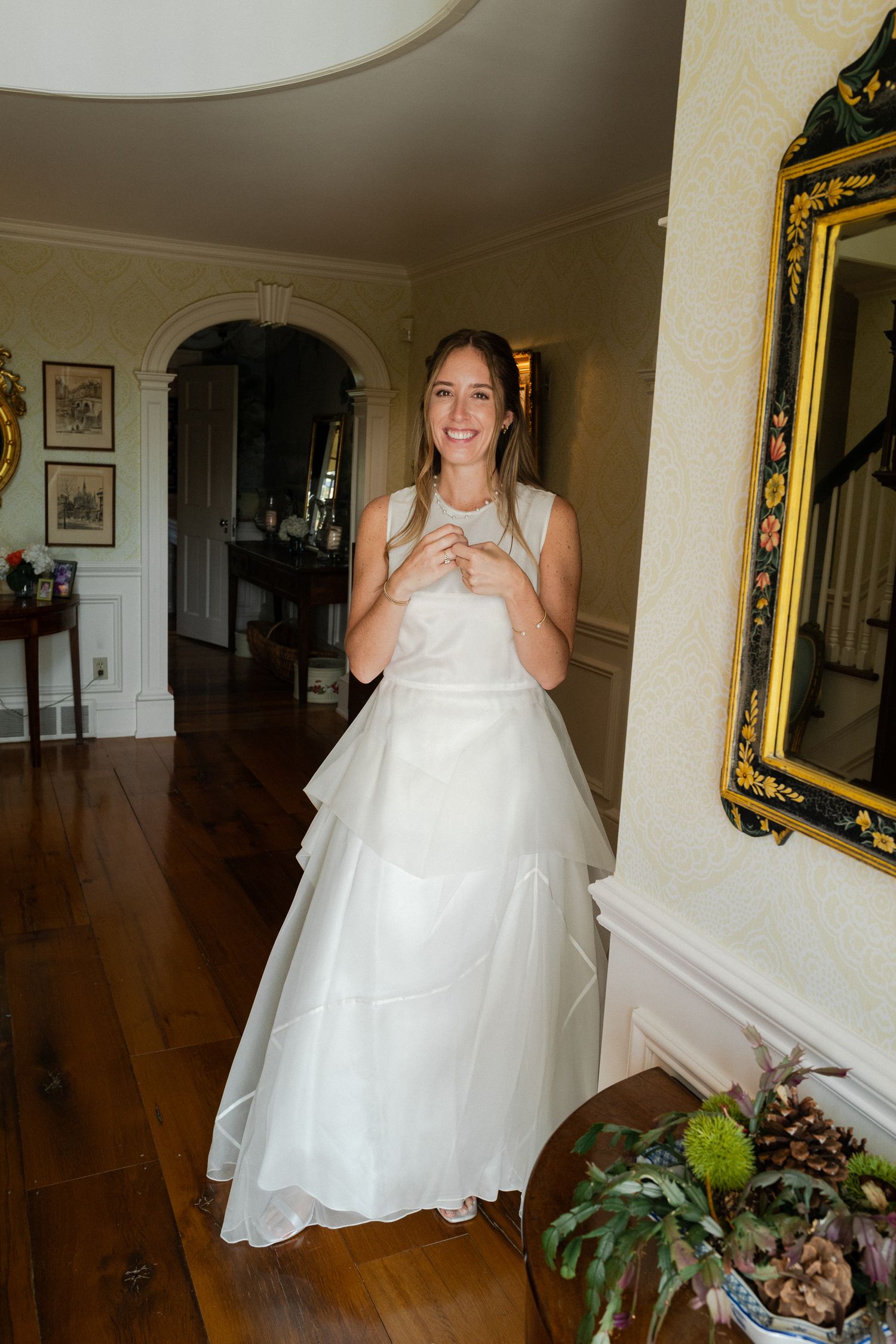 Glowing bride in white tiered wedding gown smiles in elegant home interior with ornate gold mirror.