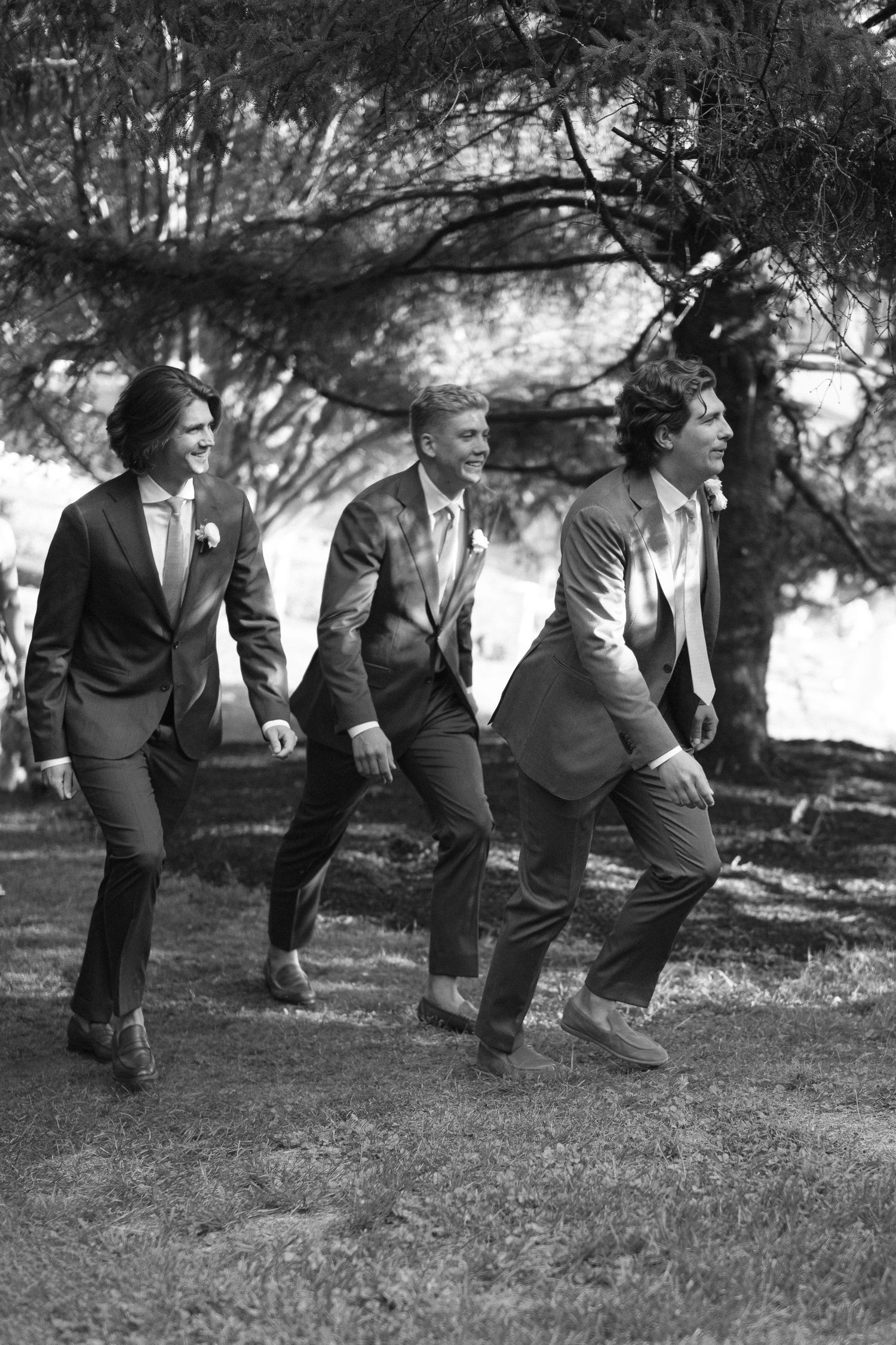 Black and white photo of three groomsmen in suits walking and laughing together under trees at a wedding.