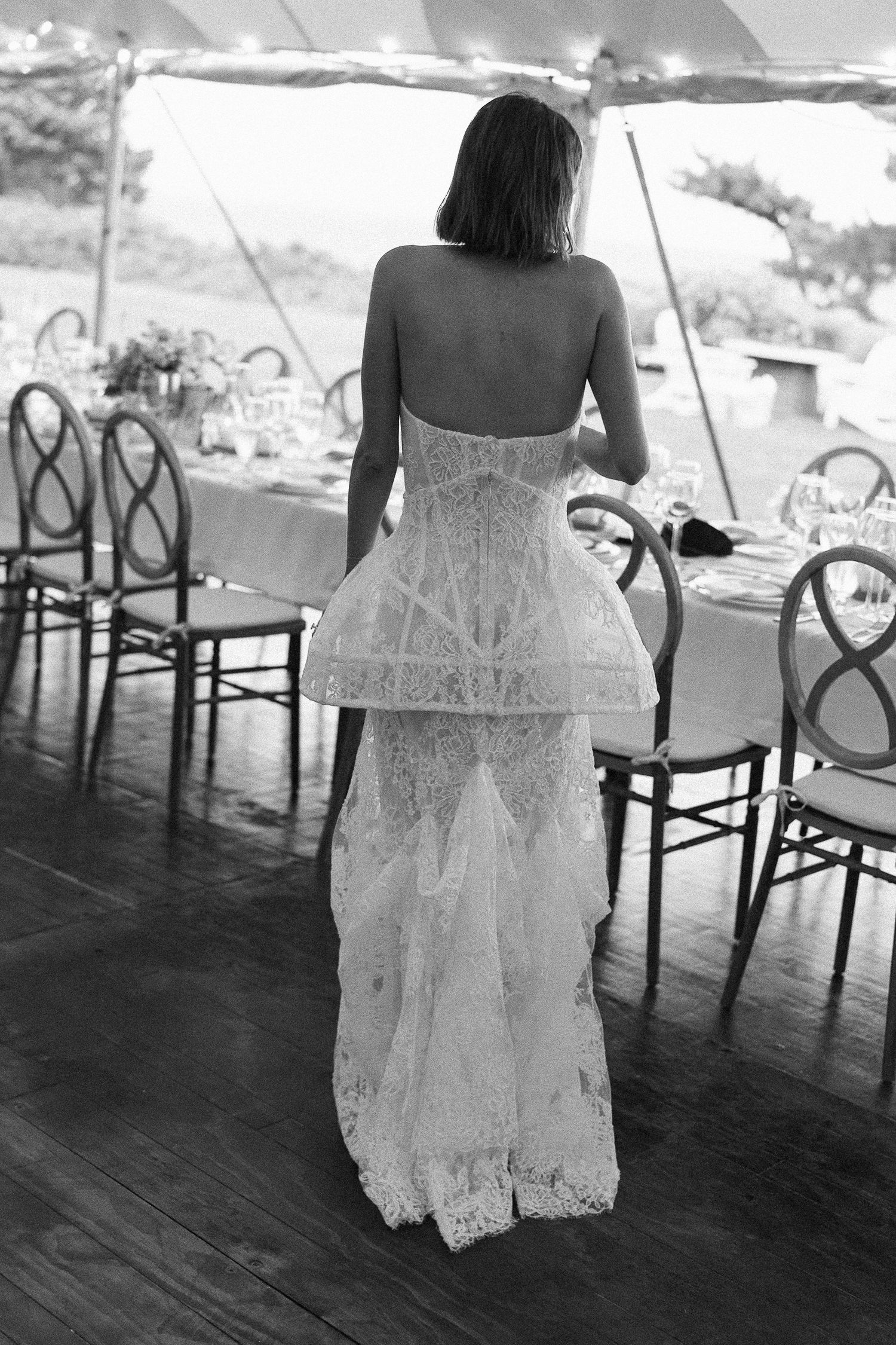 Bride in backless lace corset wedding gown standing in elegant tented reception venue, black and white photo.