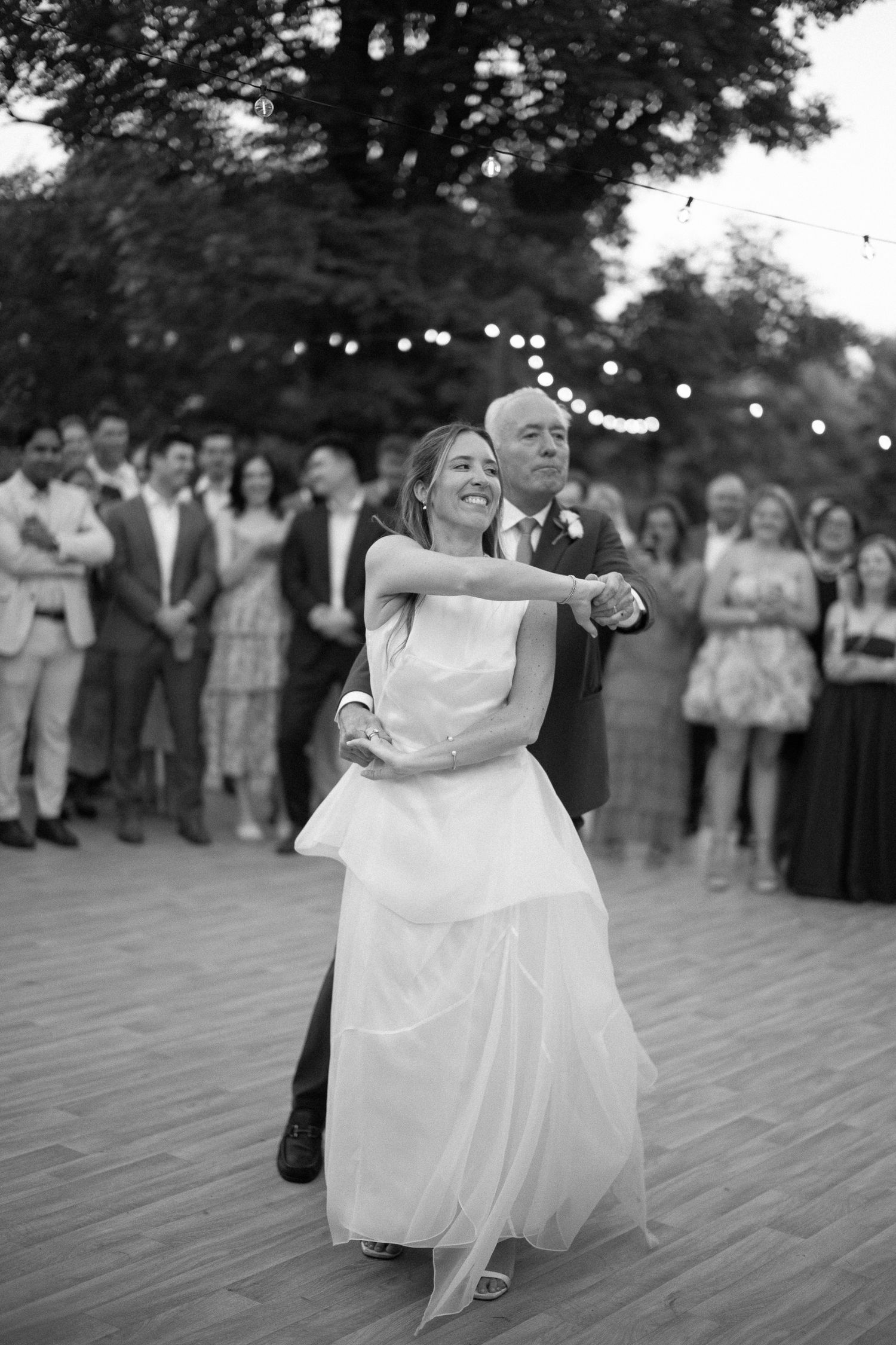Bride and father share an emotional first dance at outdoor wedding reception with string lights and guests watching.