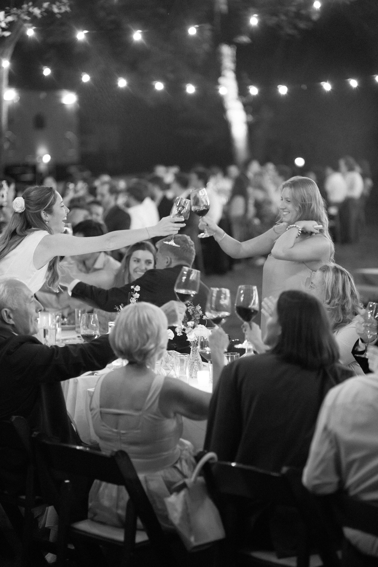 Black and white photo of wedding guests raising wine glasses in a toast under string lights at an outdoor reception.
