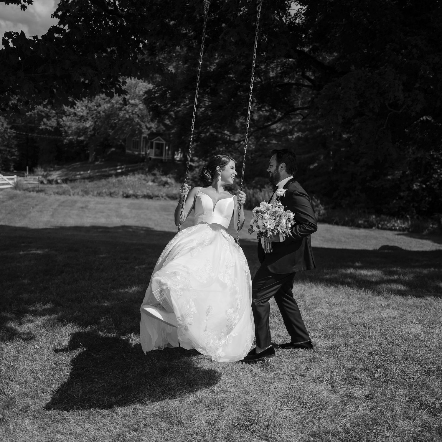 Bride sitting on a rope swing in wedding gown while groom pushes her in sunny garden.