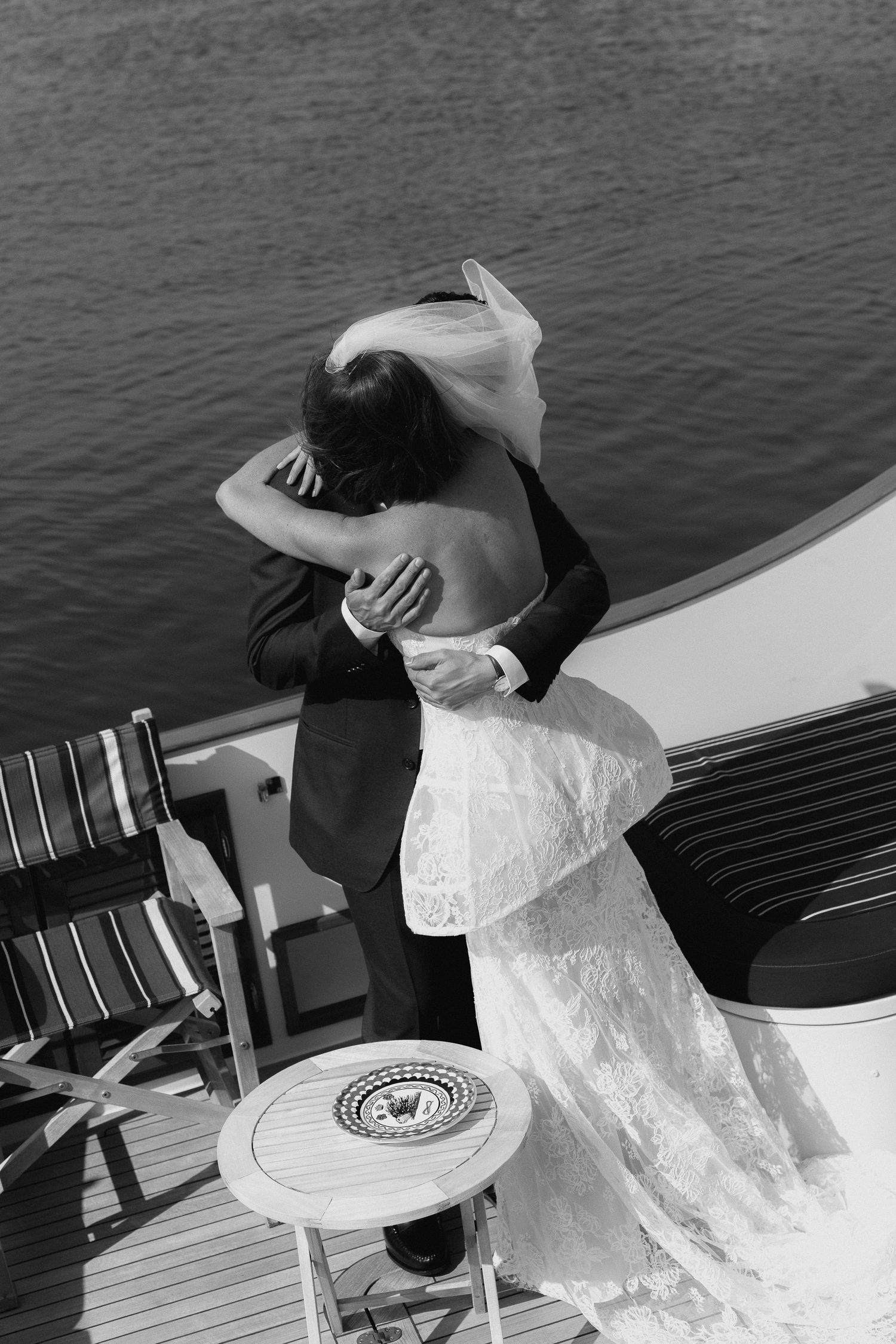 Black and white aerial view of bride and groom embracing on yacht deck above shimmering water.