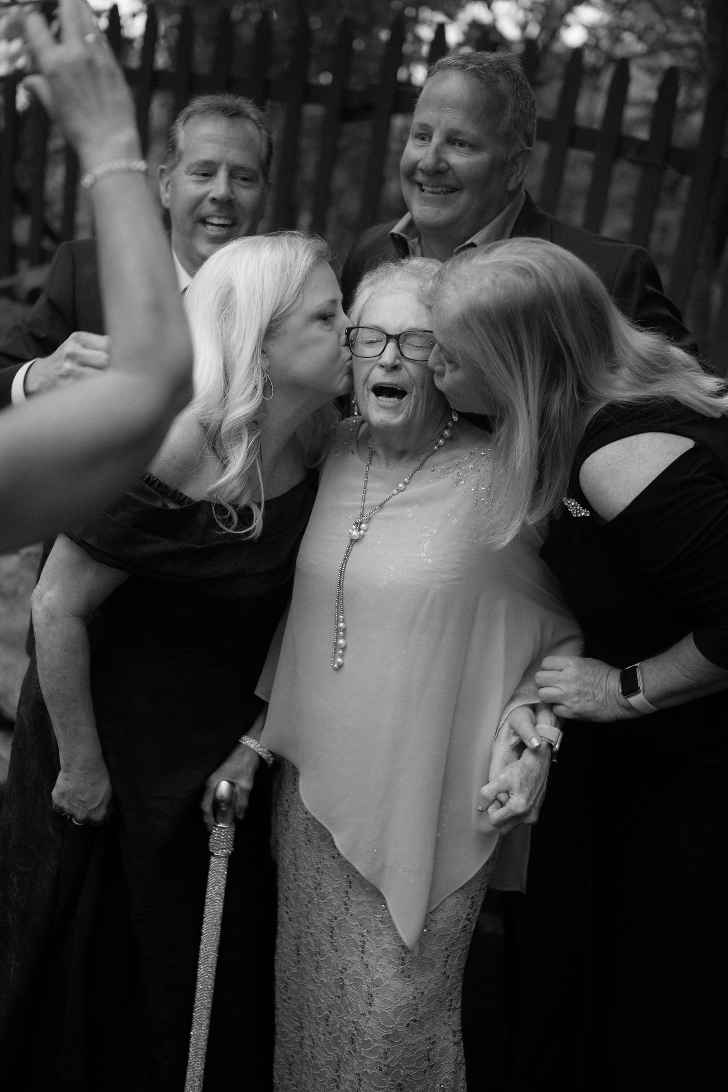 Family members kiss elderly grandmother on cheeks as men smile behind them at outdoor wedding.