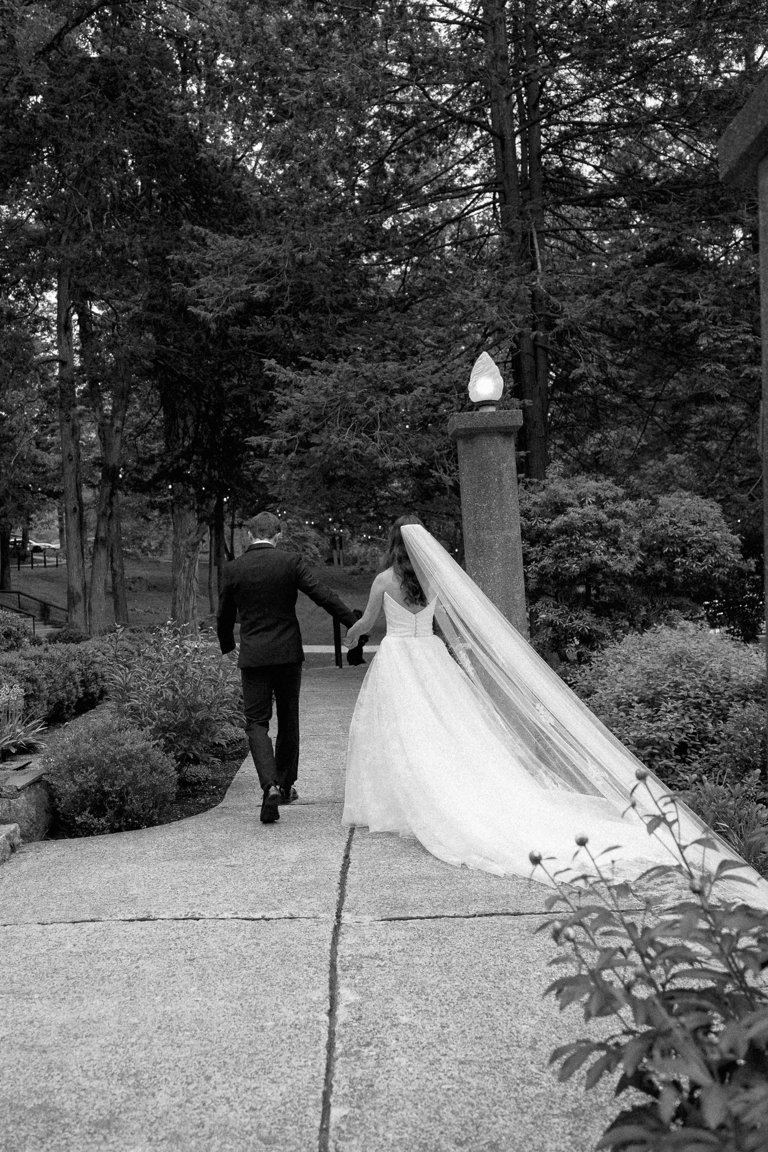 Black and white photo of newlyweds walking hand in hand through a lush garden after ceremony.