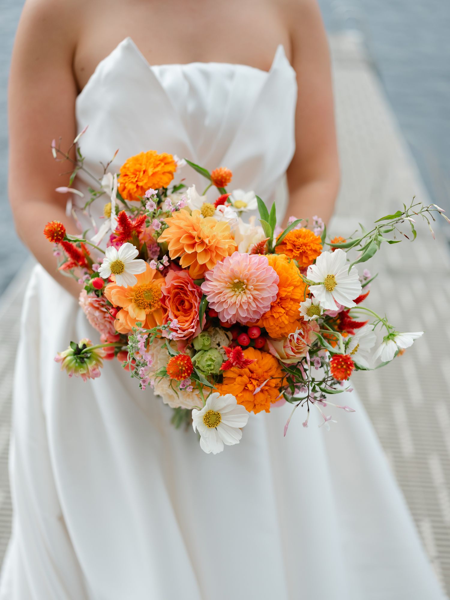 Close-up of lush bridal bouquet with orange dahlias, marigolds, cosmos, and wildflowers in white dress.