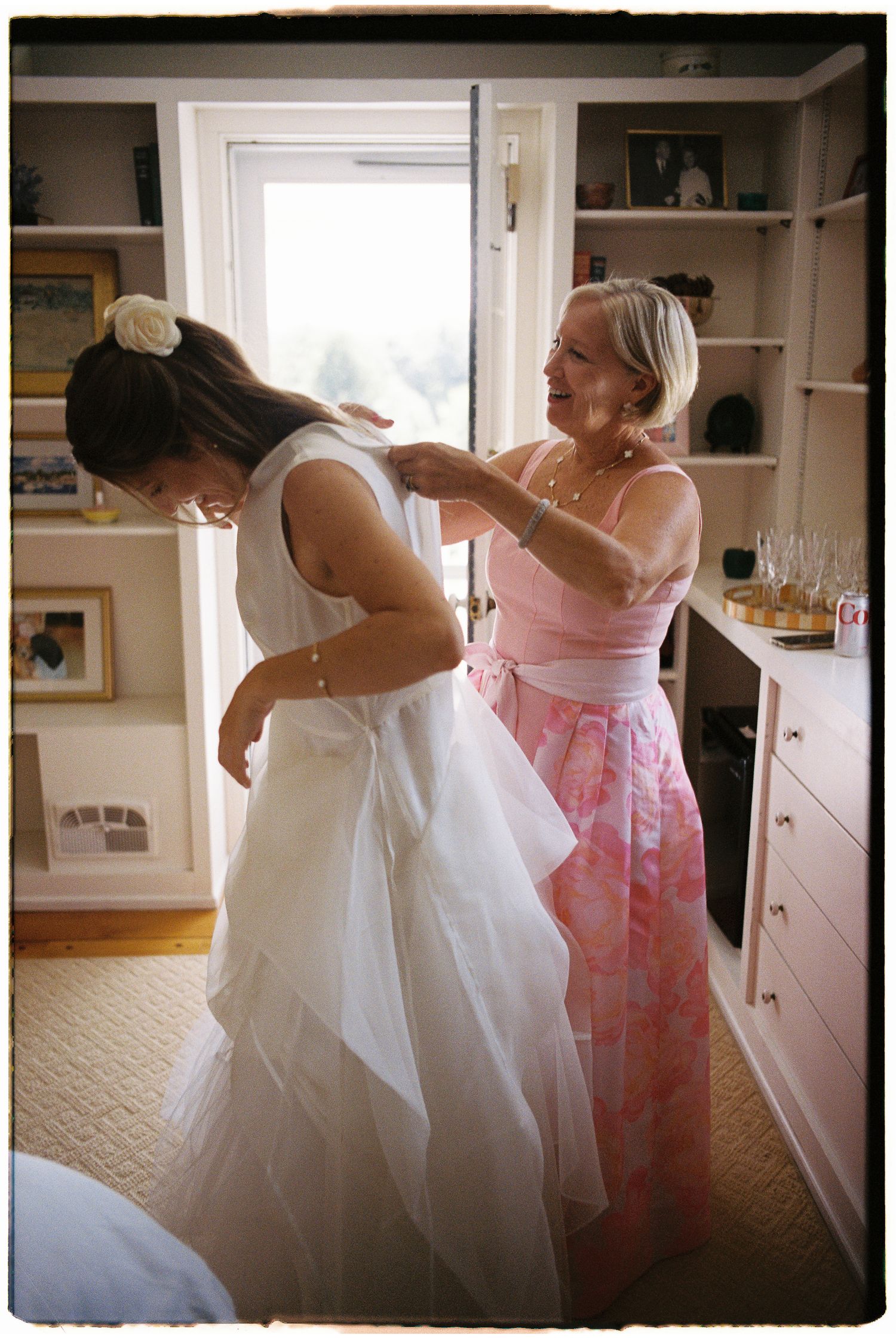An older woman in pink helps a bride button her white wedding gown before the ceremony.