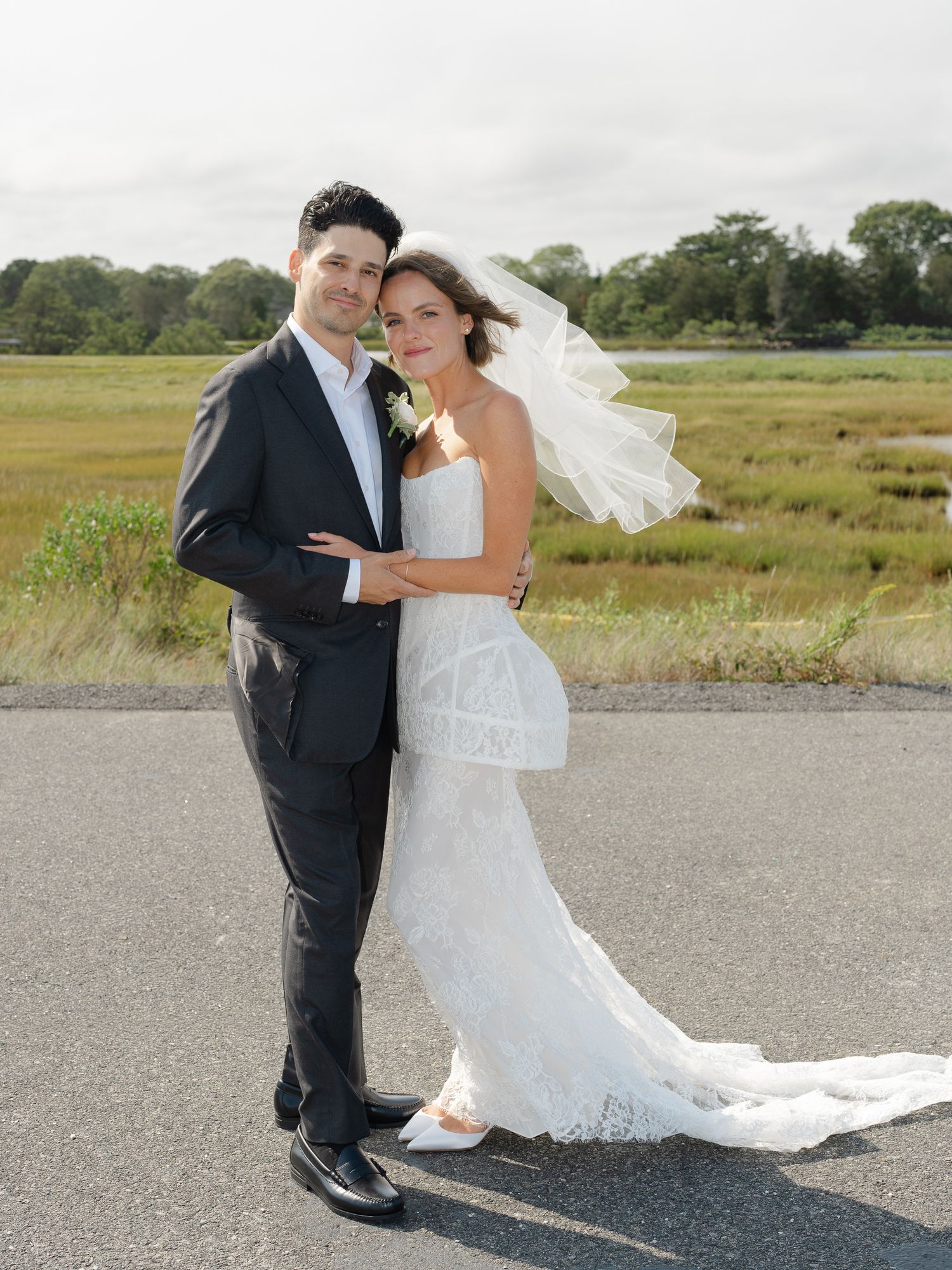 Bride and groom pose together on road with marshland backdrop, veil flowing in the breeze.