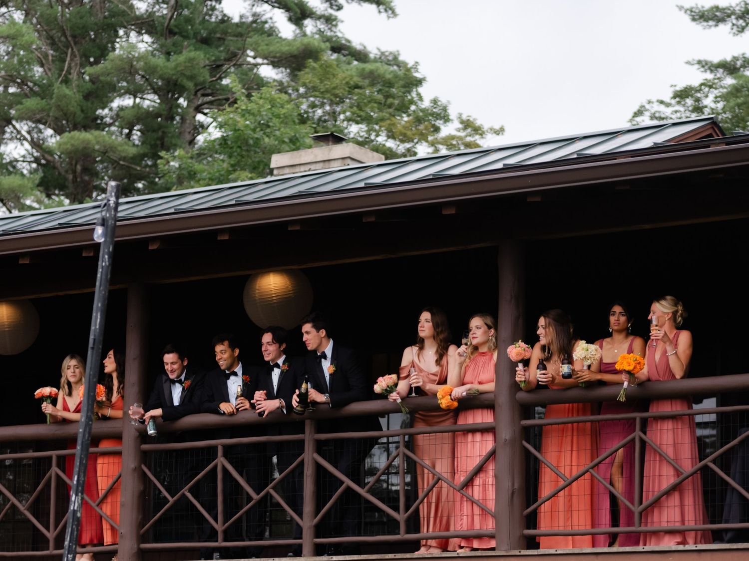 Wedding party in coral dresses and tuxedos gathered on a wooden balcony surrounded by tall trees.