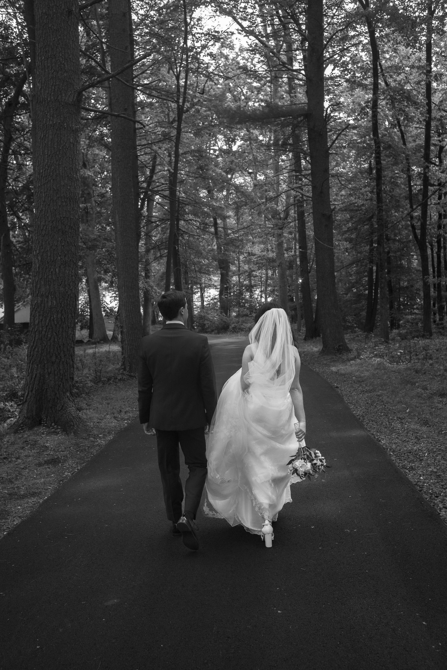 Bride and groom walk away down tree-lined path, bridal veil flowing in the breeze.