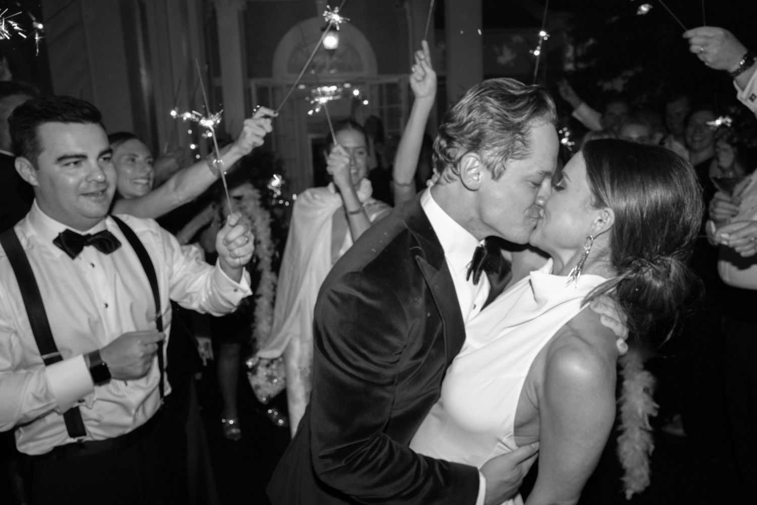 Bride and groom sharing kiss surrounded by sparklers and guests at wedding sendoff.