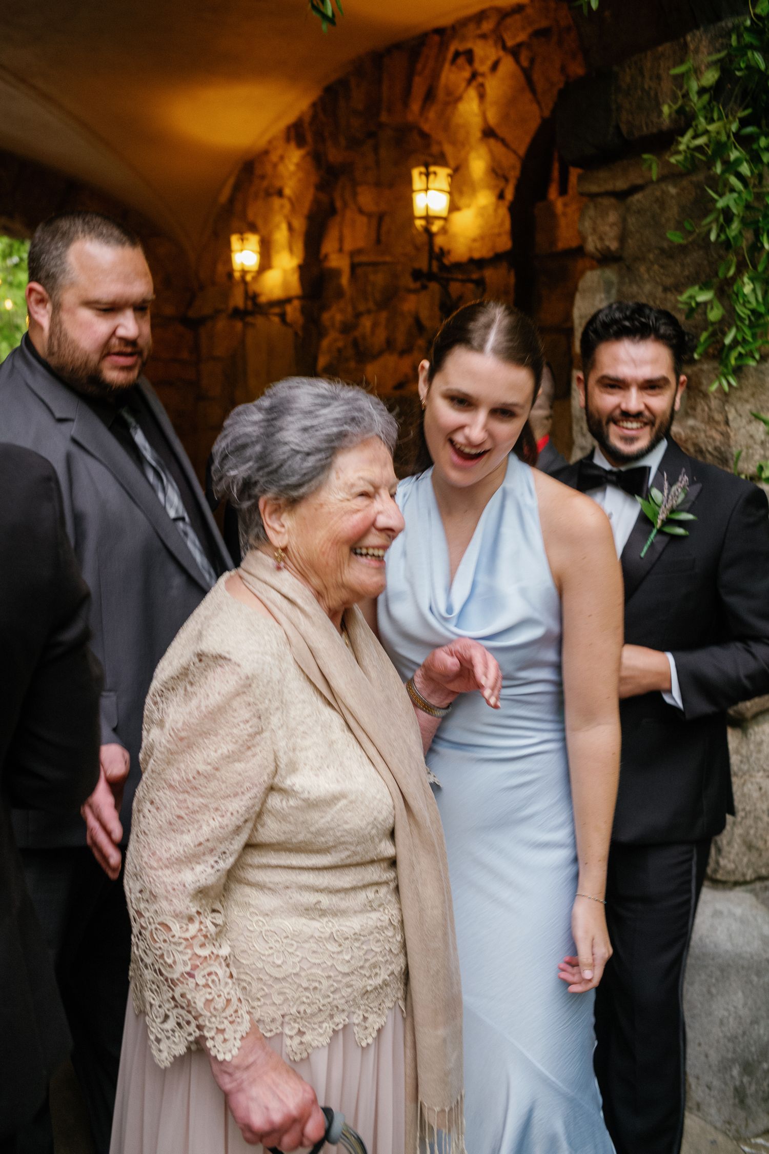 Elderly woman laughing with bridesmaid in light blue gown at elegant stone wedding venue.
