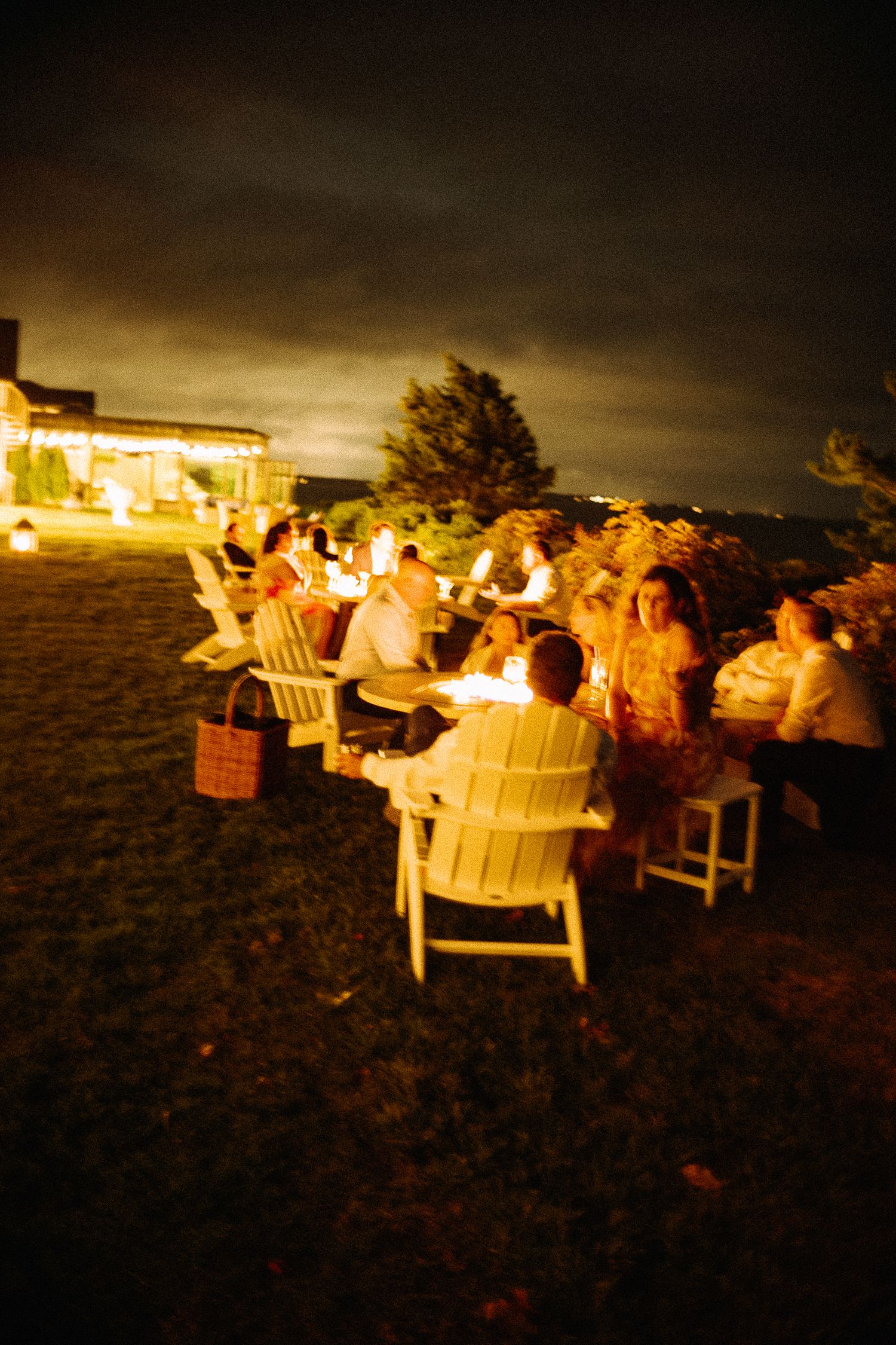 Guests gather outdoors at night around candlelit tables in white Adirondack chairs for an elegant evening party.