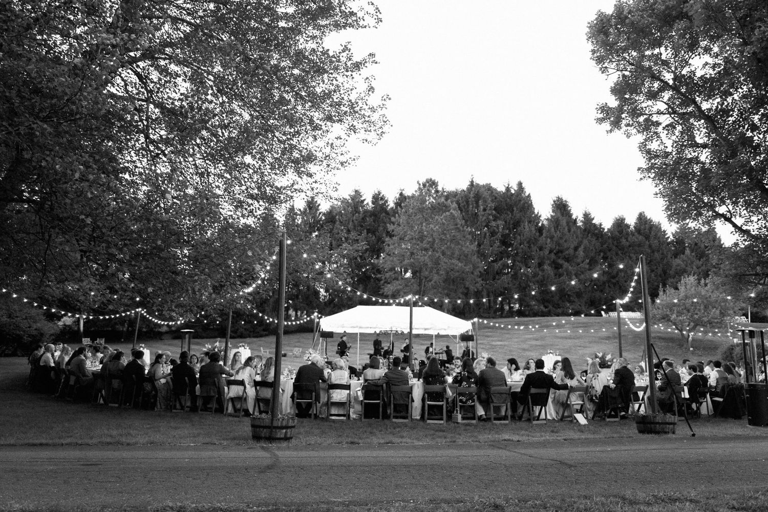 Black and white outdoor wedding reception under string lights and white tent surrounded by trees at dusk.