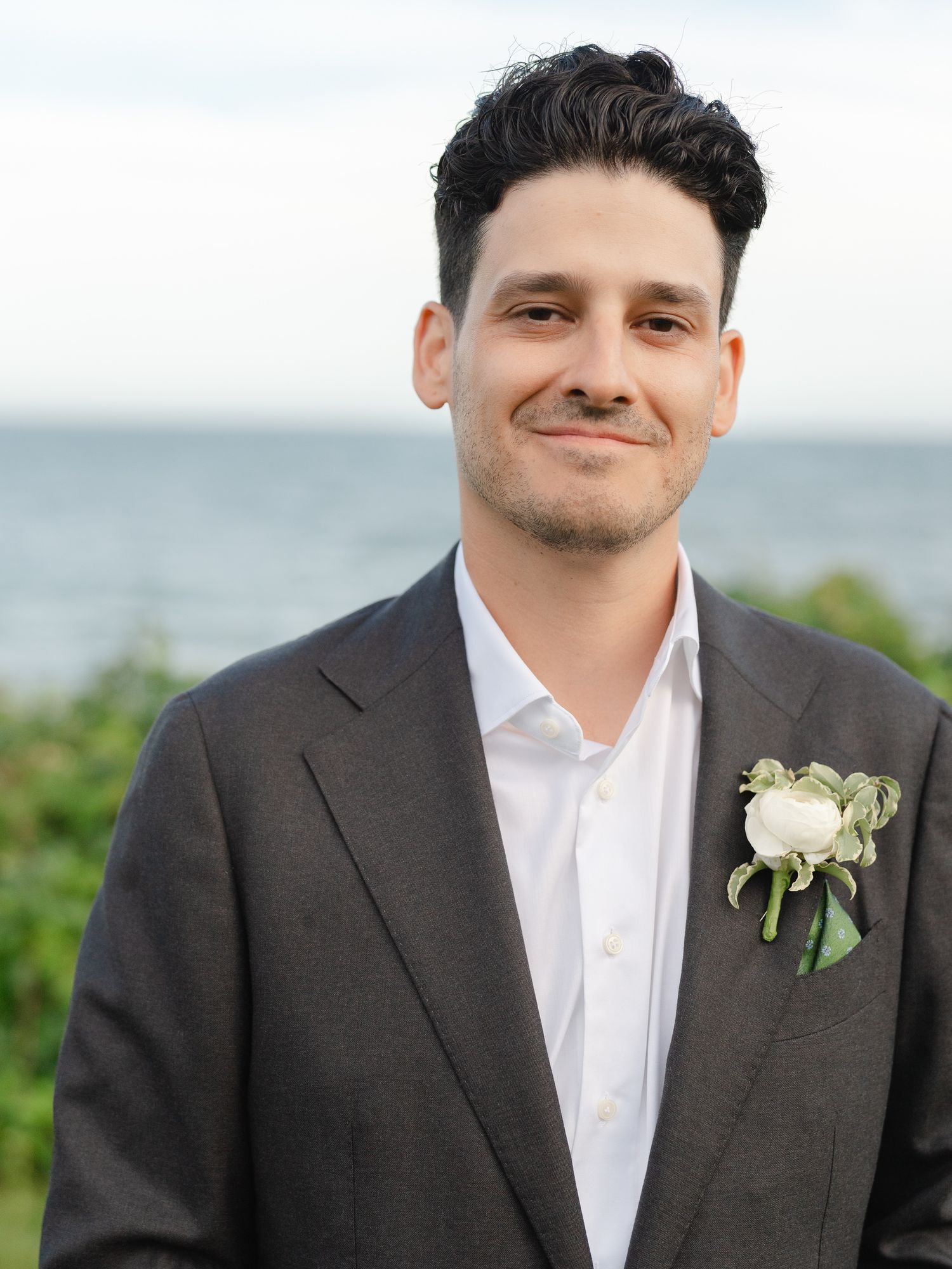 Smiling groom in dark suit with white boutonniere posing outdoors by the ocean.