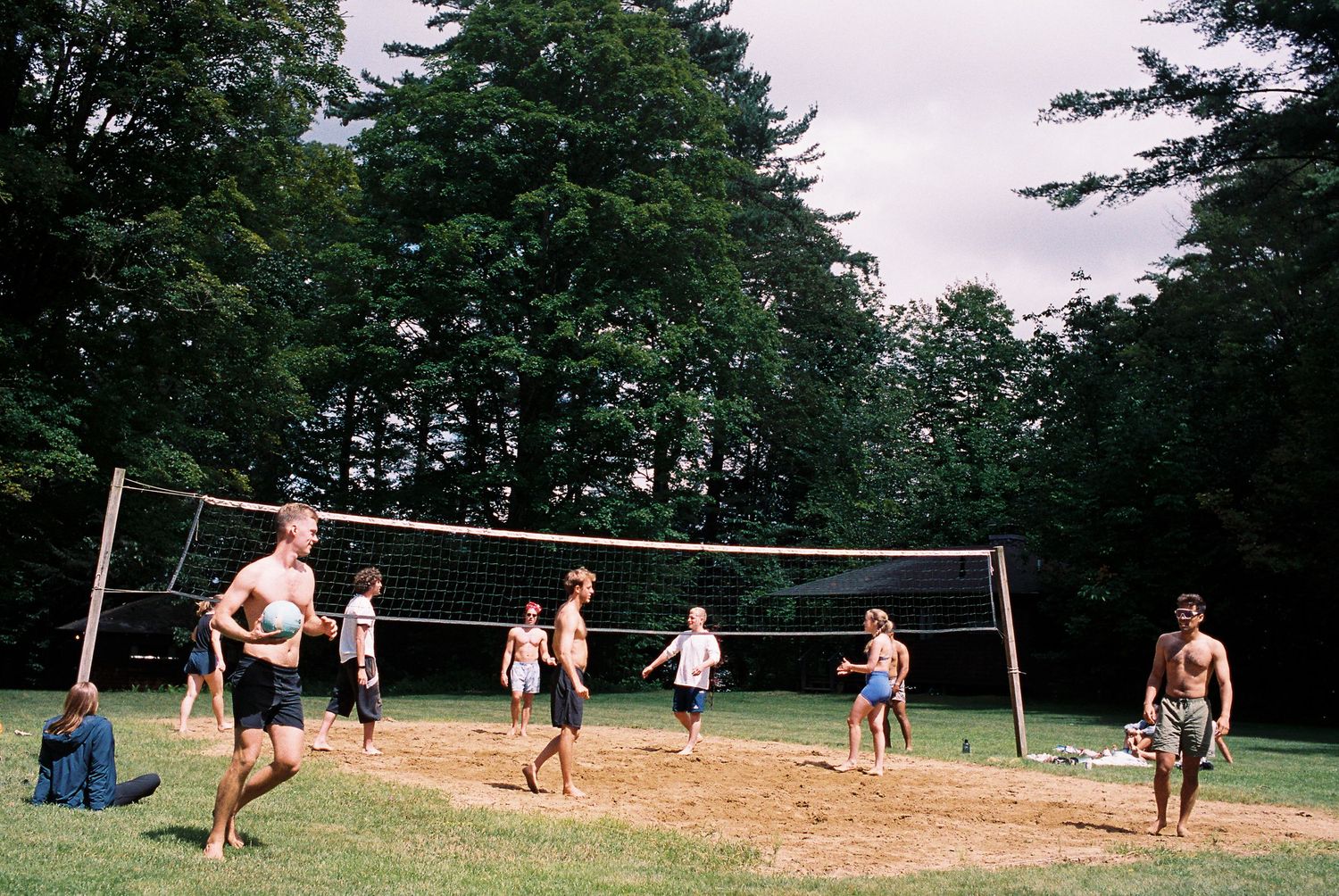 A group of guests enjoy a casual volleyball game on a sandy court surrounded by tall trees.