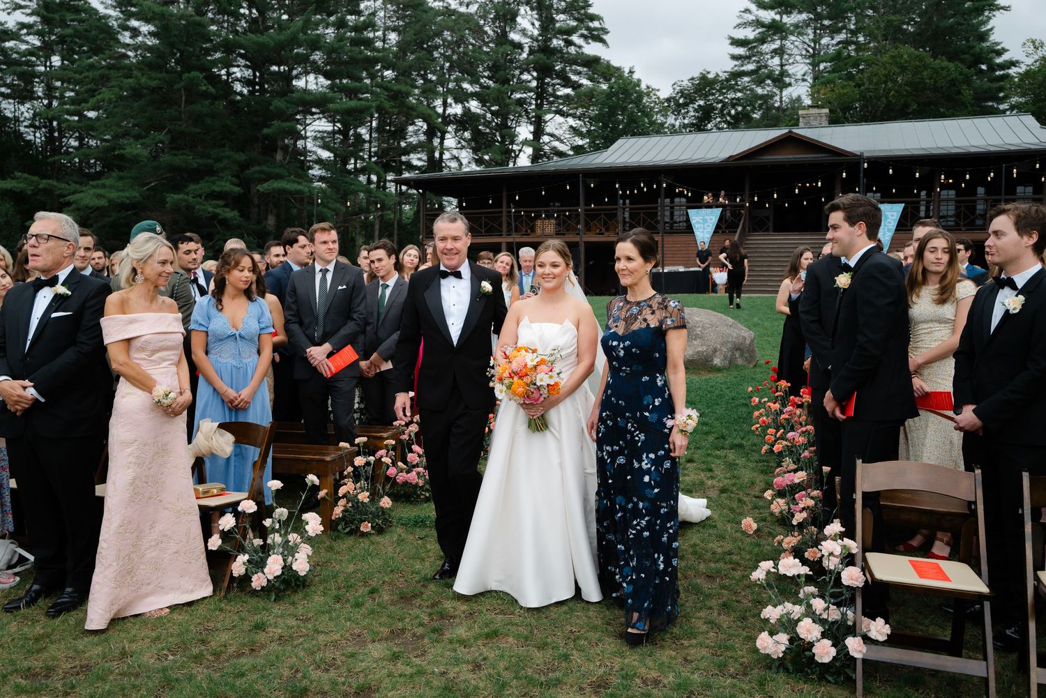 Bride in white gown walking down outdoor aisle with family, bridesmaid in navy sequin dress nearby.