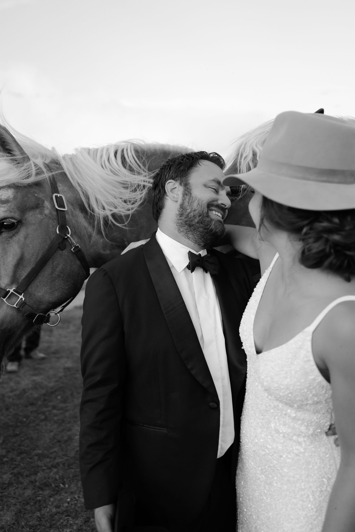 Smiling groom in tuxedo stands beside his bride near palomino horses at a rustic outdoor wedding.