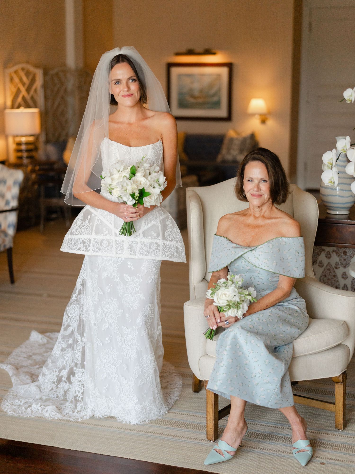 Bride in strapless lace wedding gown with veil stands beside seated older woman in silver dress.