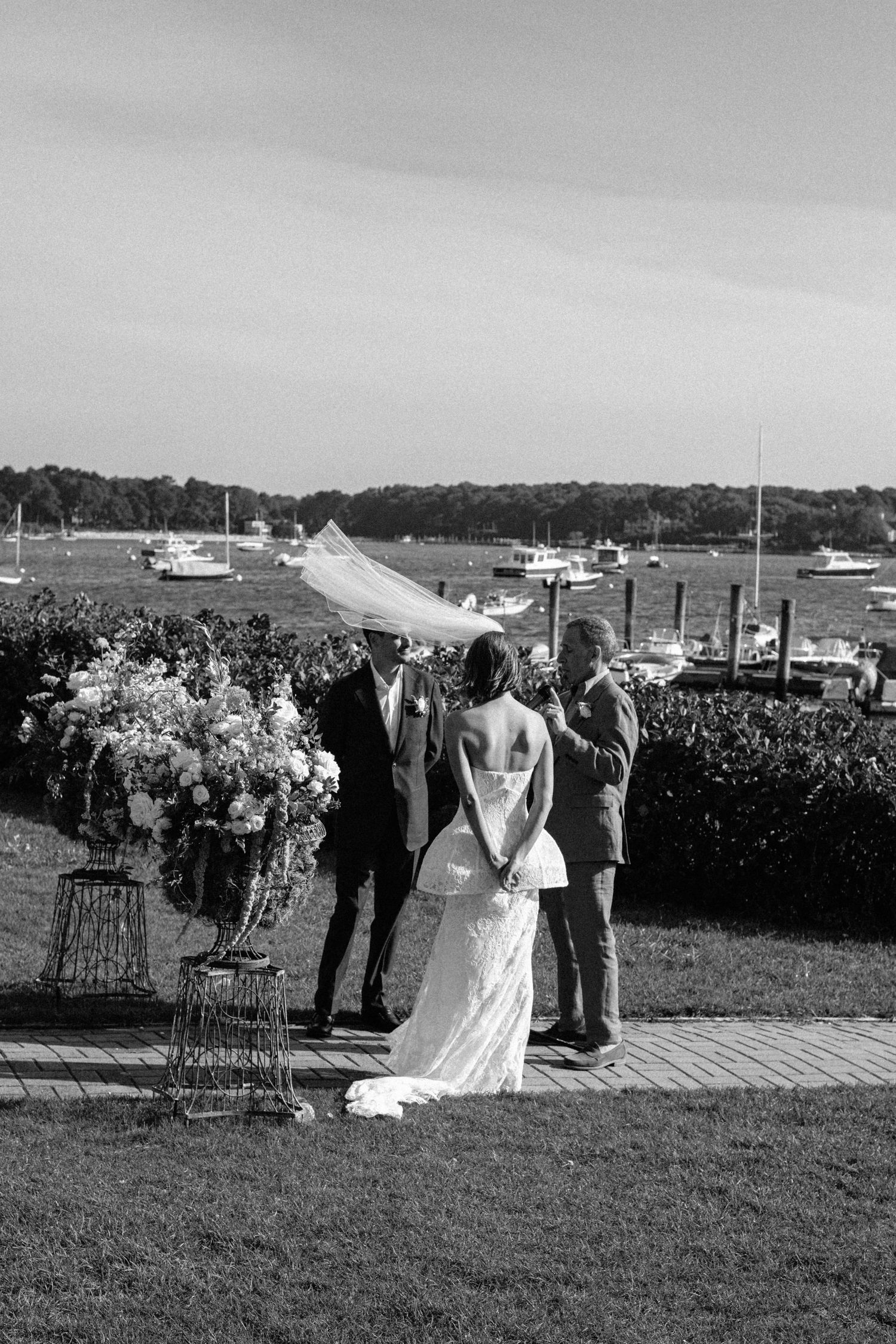 Black and white photo of bride and groom exchanging vows at waterfront ceremony with veil blowing in wind.