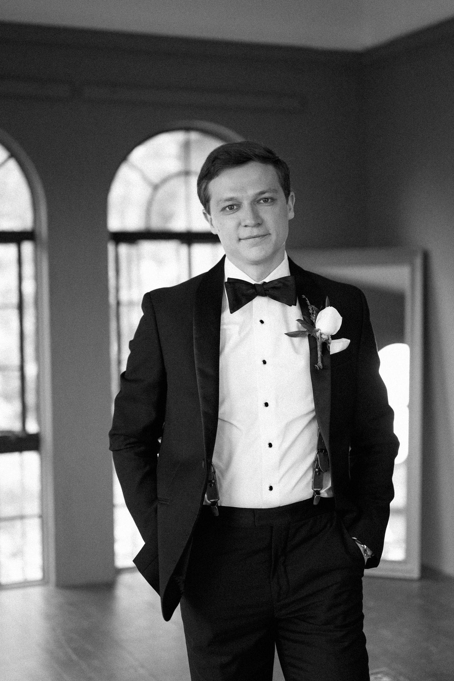 Groom in black tuxedo with bow tie and boutonniere poses in elegant room with arched windows.