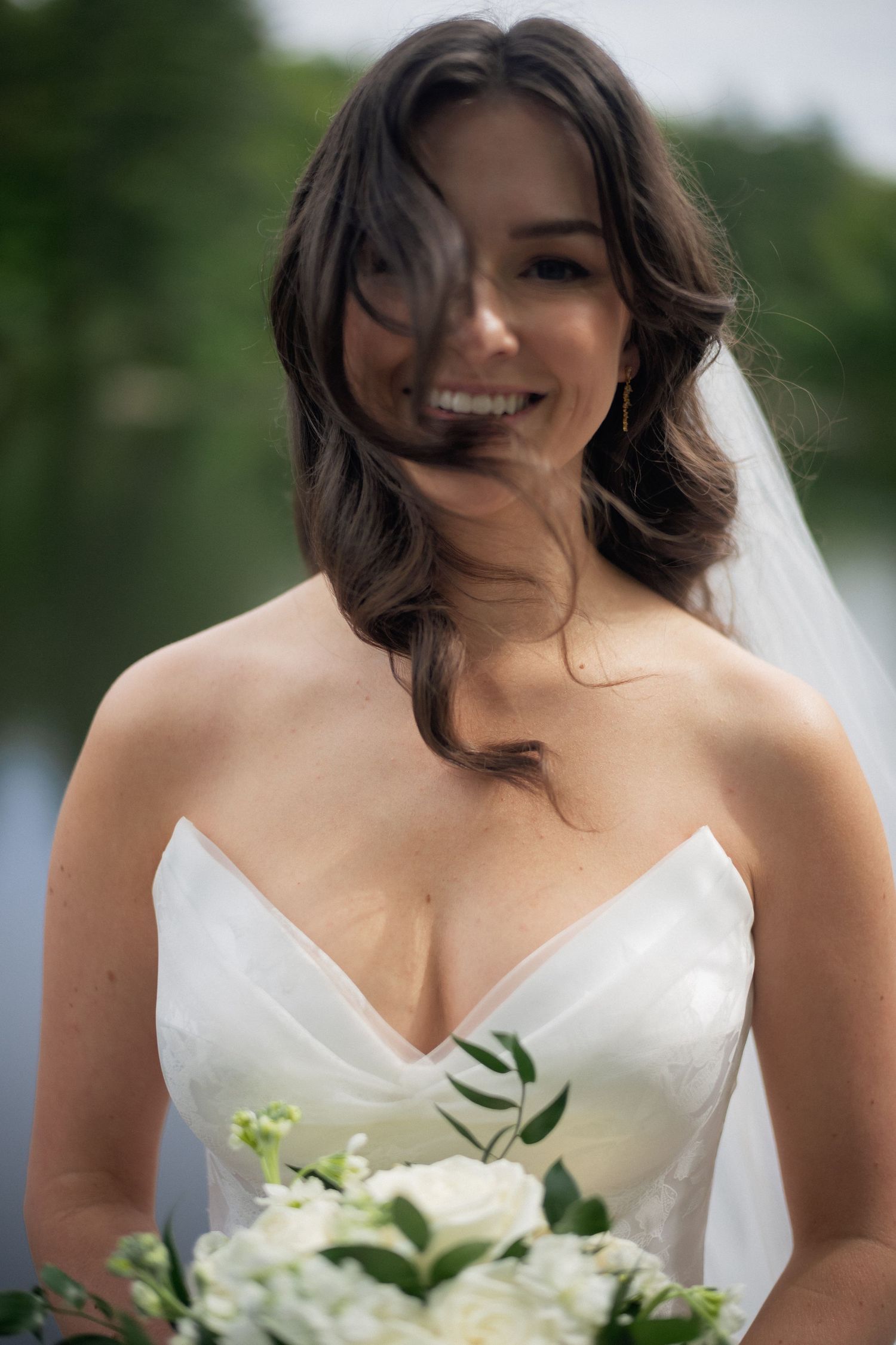 Smiling bride with wind-blown hair holding a white floral bouquet near a lake on her wedding day.