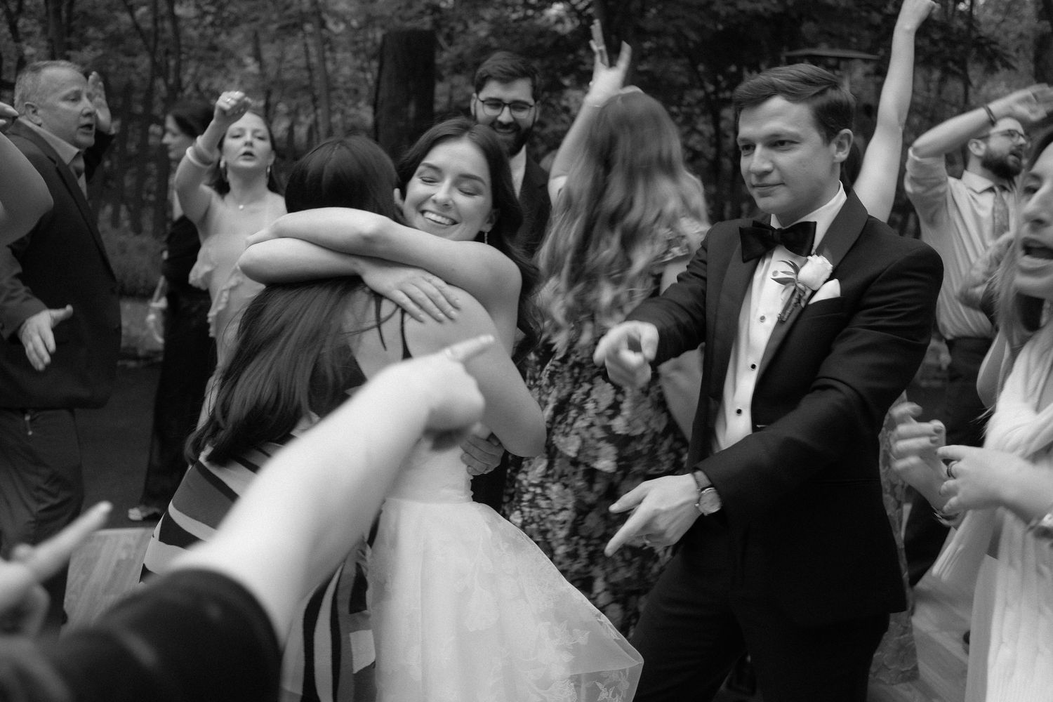 Black and white photo of a bride hugging a guest while the groom celebrates on the dance floor at a wedding reception.