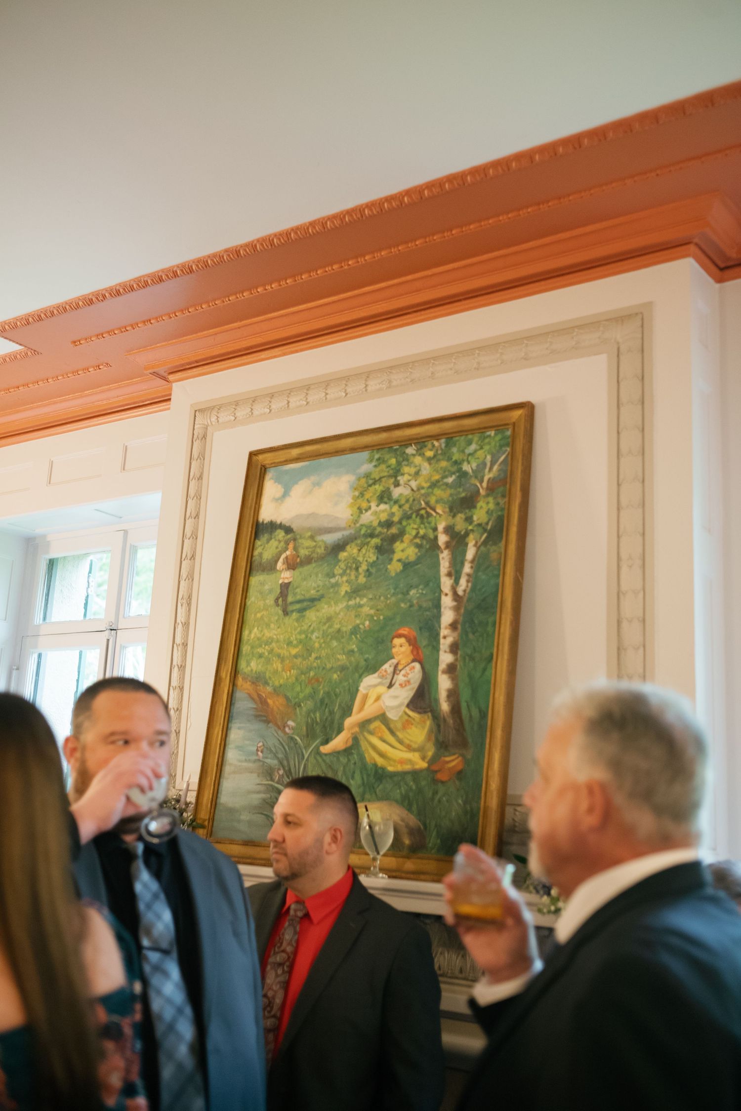 Wedding guests mingle beneath ornate painting in elegant wood-trimmed reception venue room.