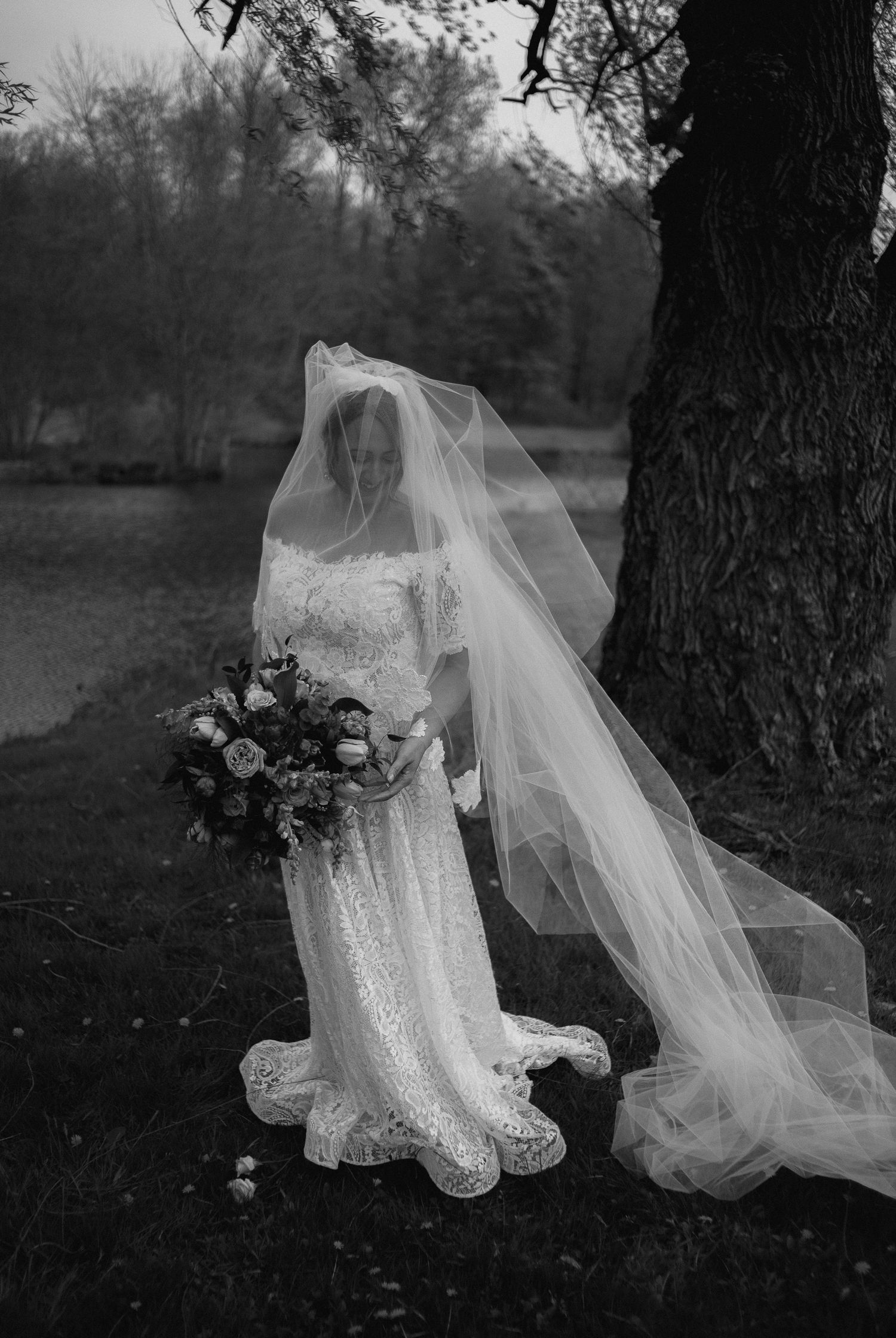 Black and white bridal portrait of a bride in lace gown with flowing veil holding bouquet near a tree.