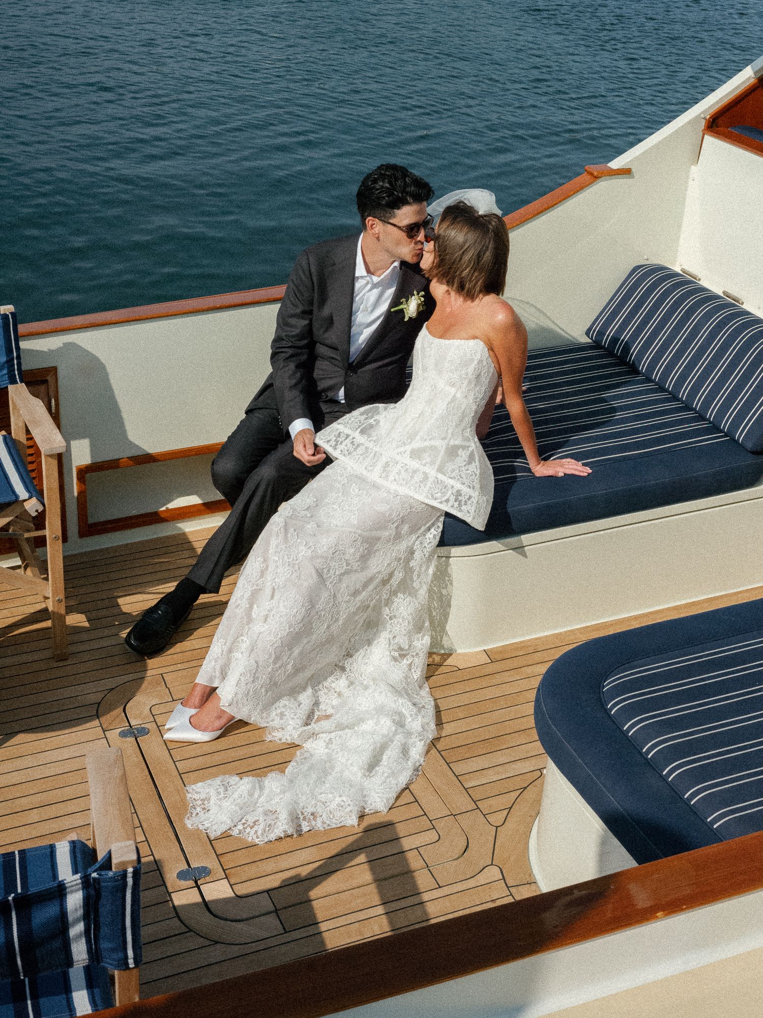 Bride and groom share romantic kiss while seated on luxury yacht deck with blue water behind them.