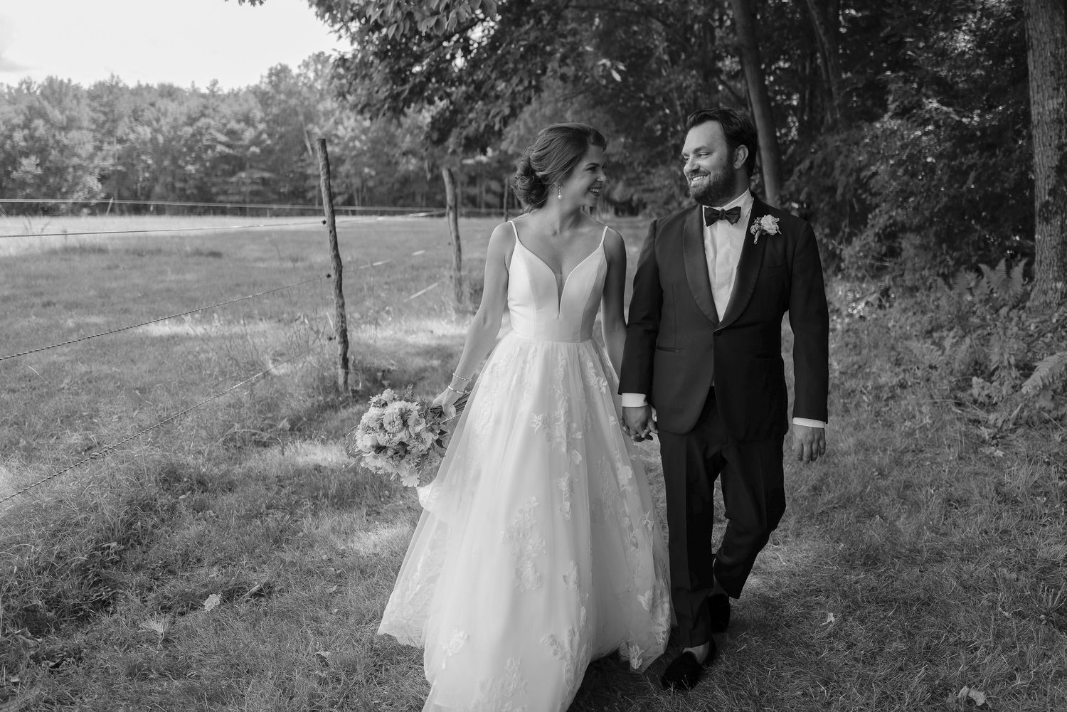 Bride and groom holding hands walking along a rustic fence with lush green trees behind them.
