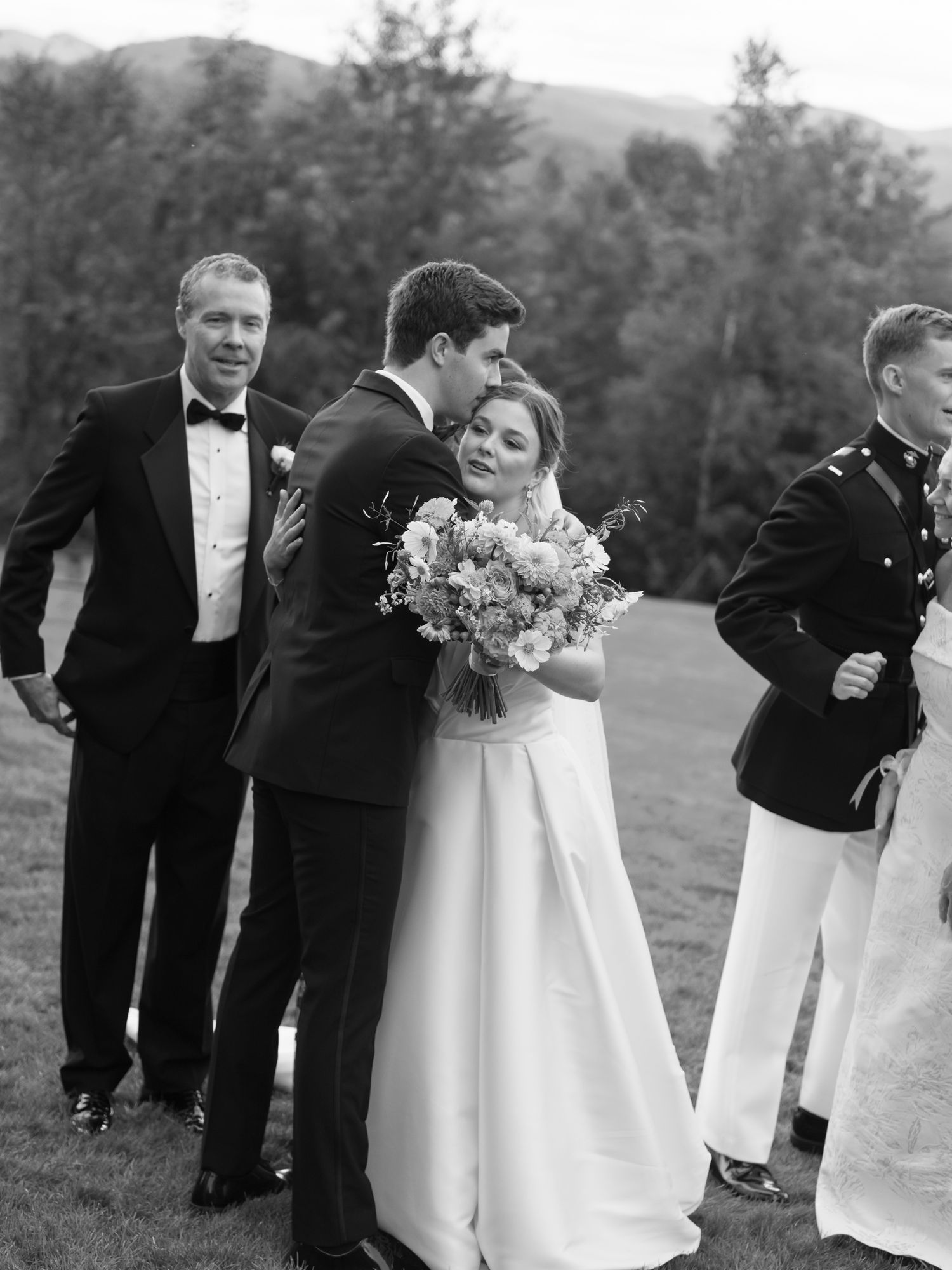 Black and white photo of bride and groom embracing at outdoor wedding ceremony with guests in formal attire.