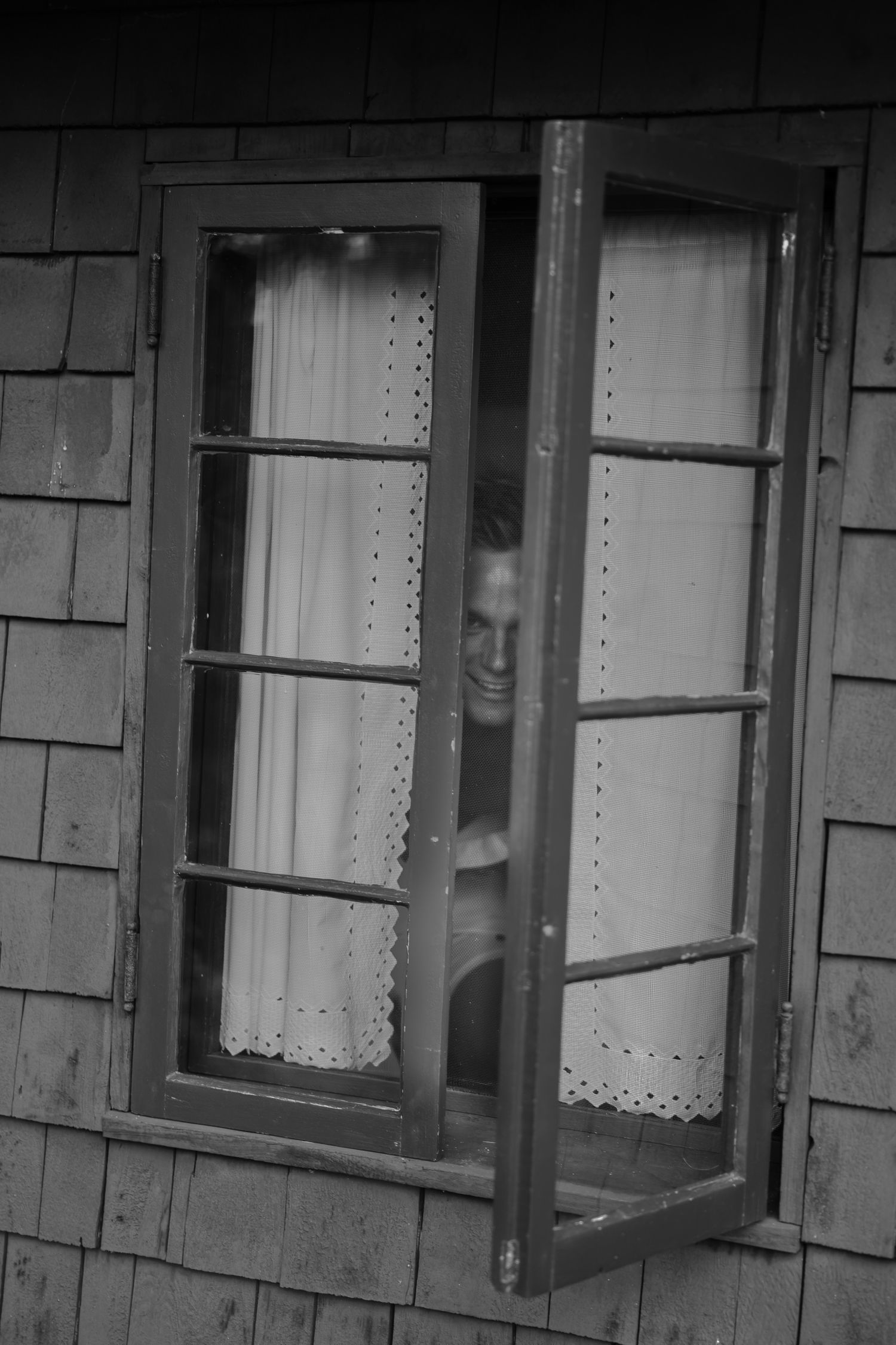 Black and white photo of a person smiling through an open vintage window of a shingled building.