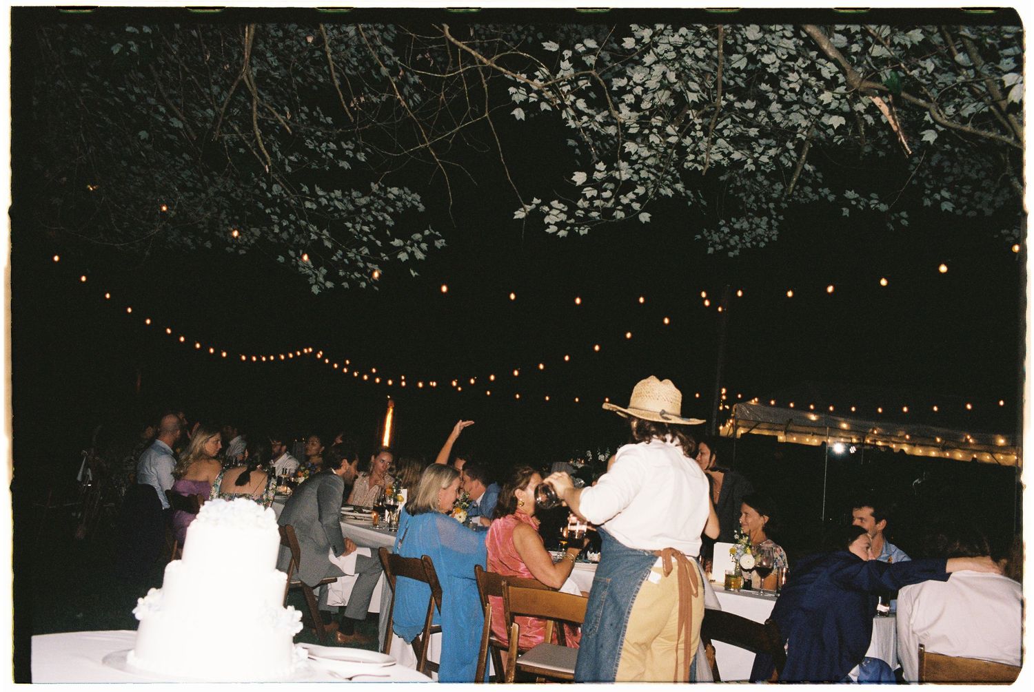 Wedding guests gather under string lights at a nighttime outdoor reception with a white tiered cake.