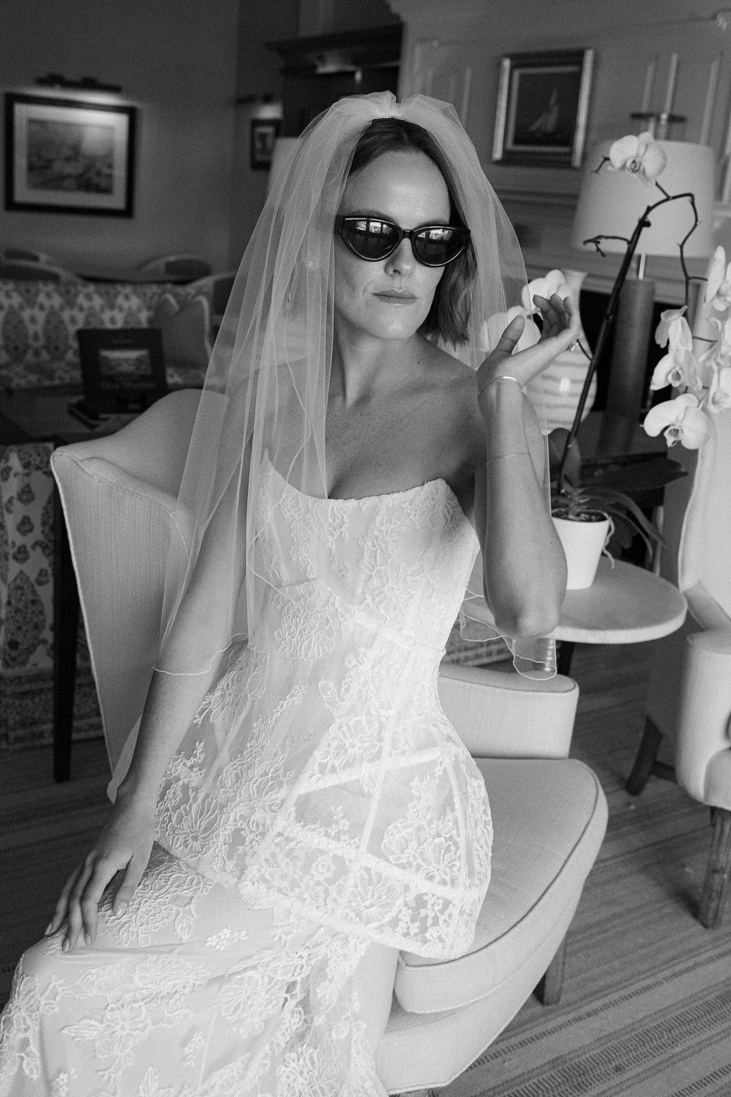 Black and white photo of a bride in lace gown and veil wearing sunglasses, seated in an elegant room.