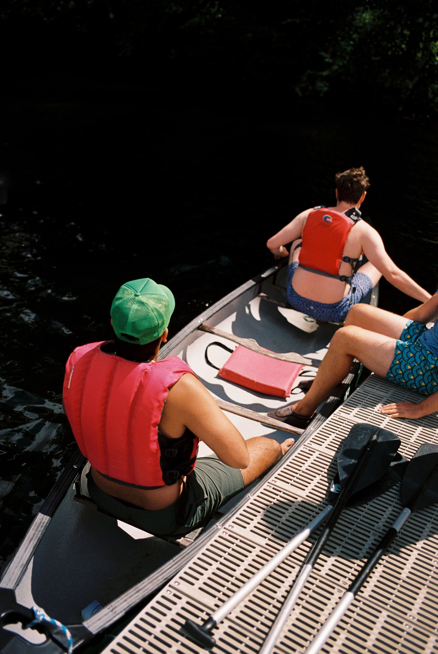 Two people wearing red life jackets paddling a canoe through dark shaded water.