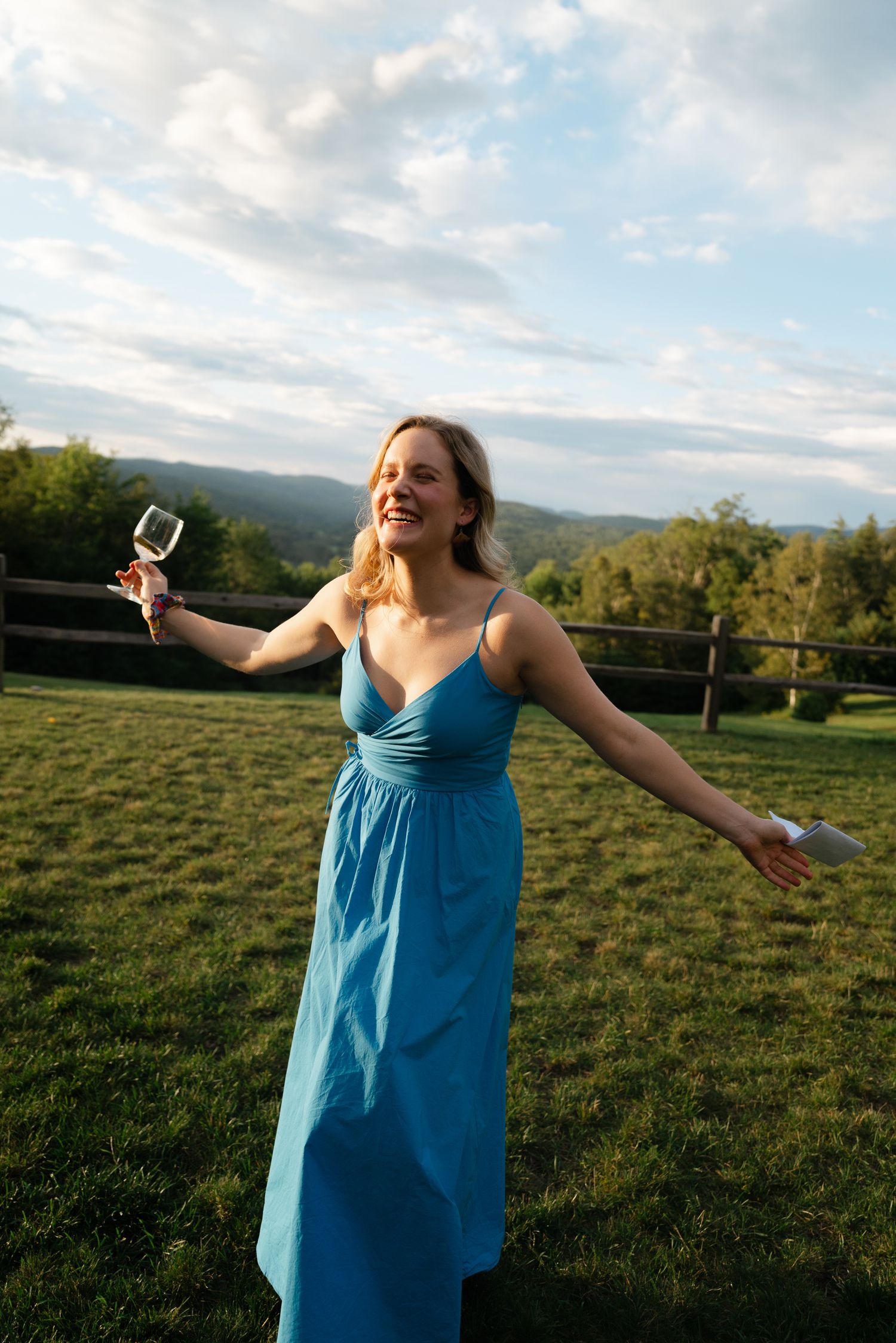 A joyful woman in a blue dress holds a wine glass with arms outstretched on a scenic green hillside.