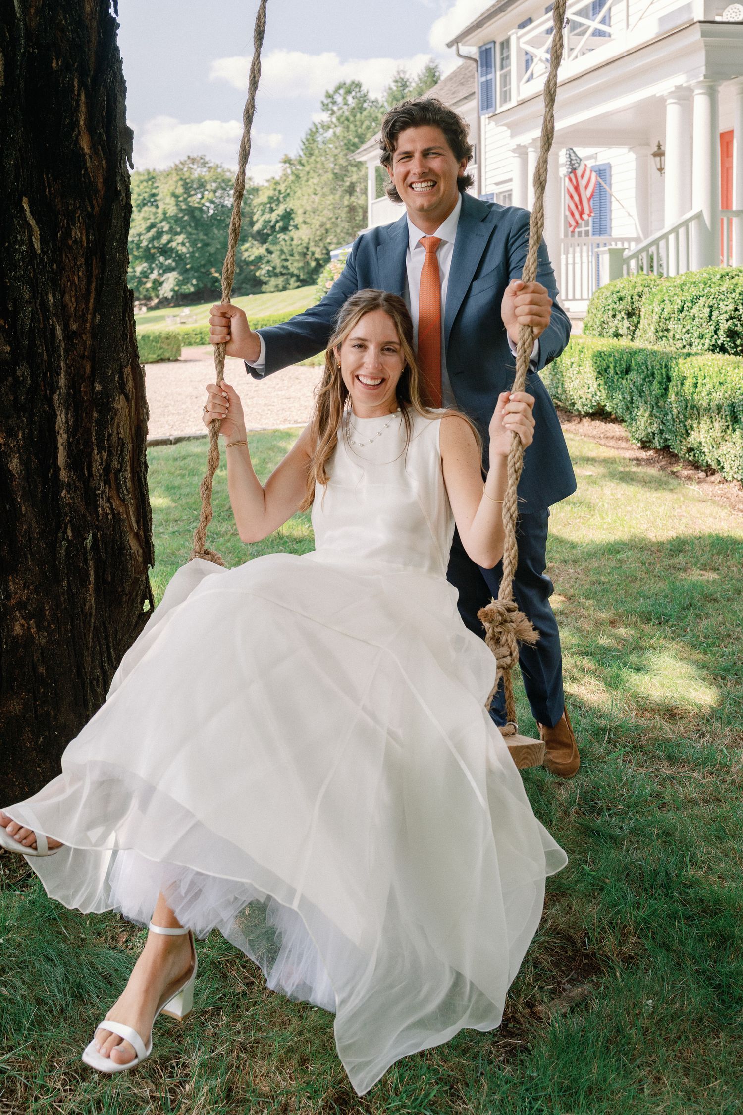 Happy bride sitting on a rope swing as groom pushes her outside a colonial home.