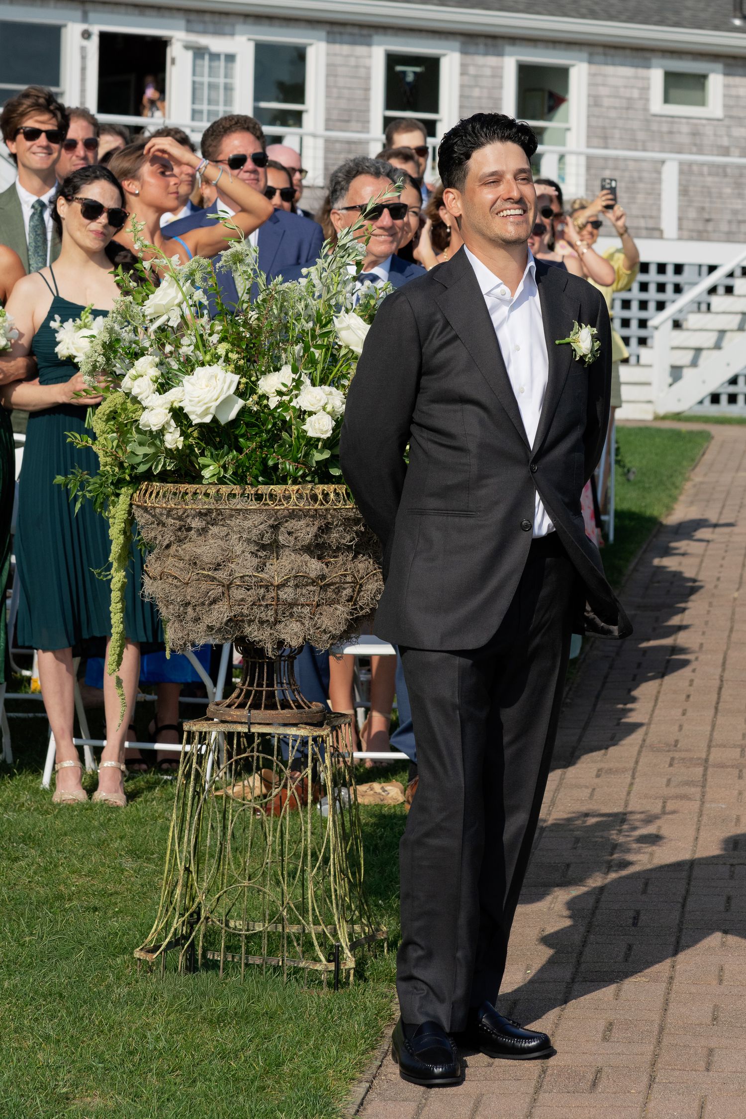 Smiling groom in dark suit standing near floral arrangement at outdoor wedding ceremony.