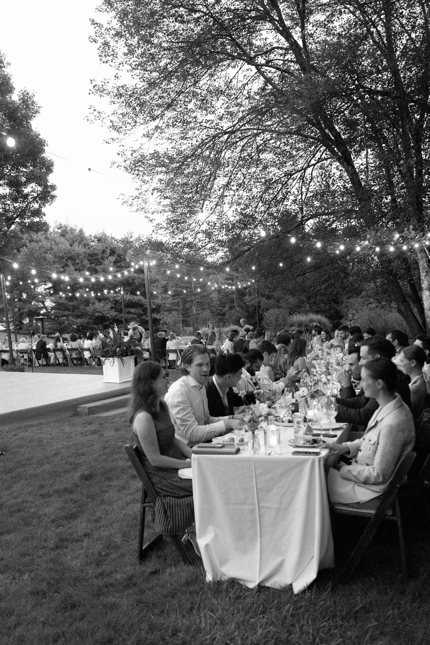Guests seated at long elegantly set dinner tables under string lights at outdoor wedding reception at dusk.