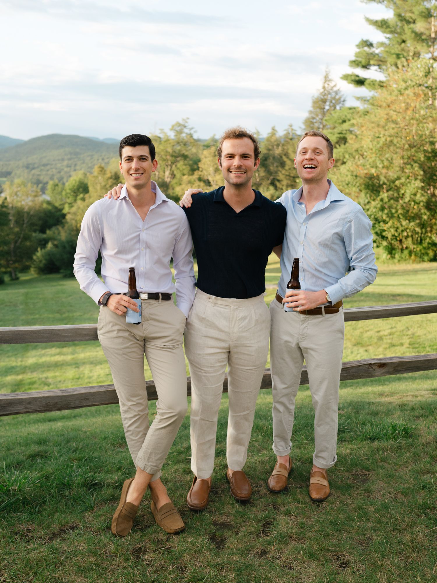 Three smiling men in smart casual attire pose together holding beers at an outdoor wedding event.