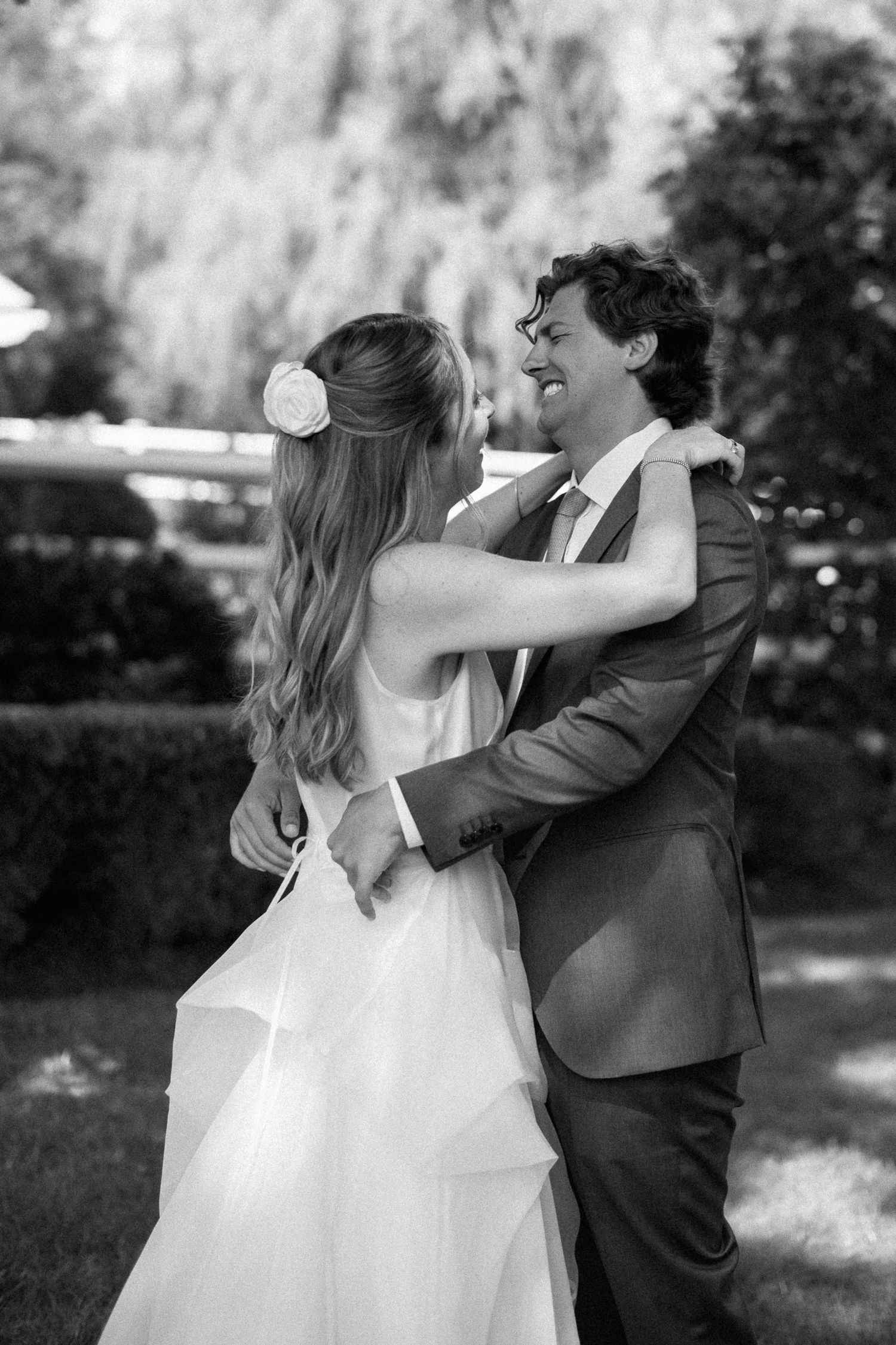 Black and white photo of smiling bride and groom embracing outdoors in a garden.