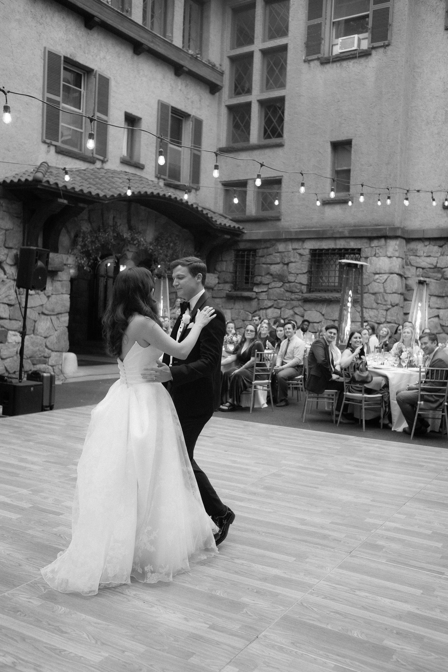Bride and groom share their first dance at an outdoor wedding reception in a stone courtyard with string lights.